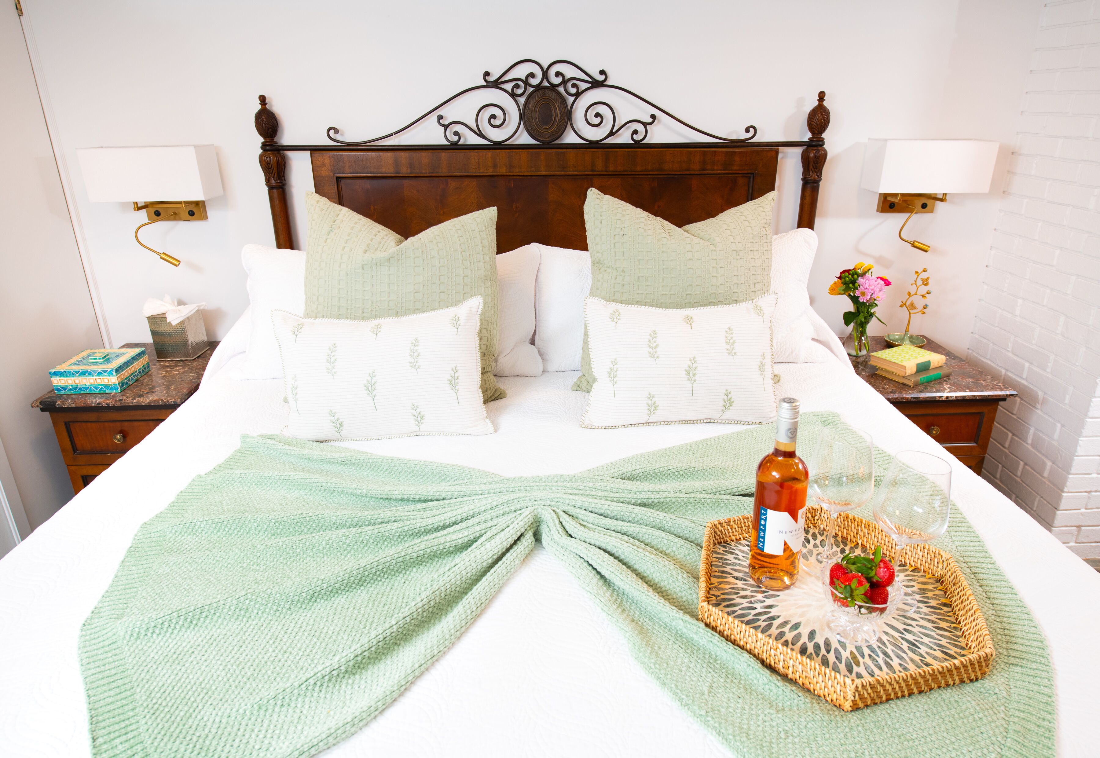 A neatly made bed with a green blanket, a bottle of wine, and a fruit basket on a tray.