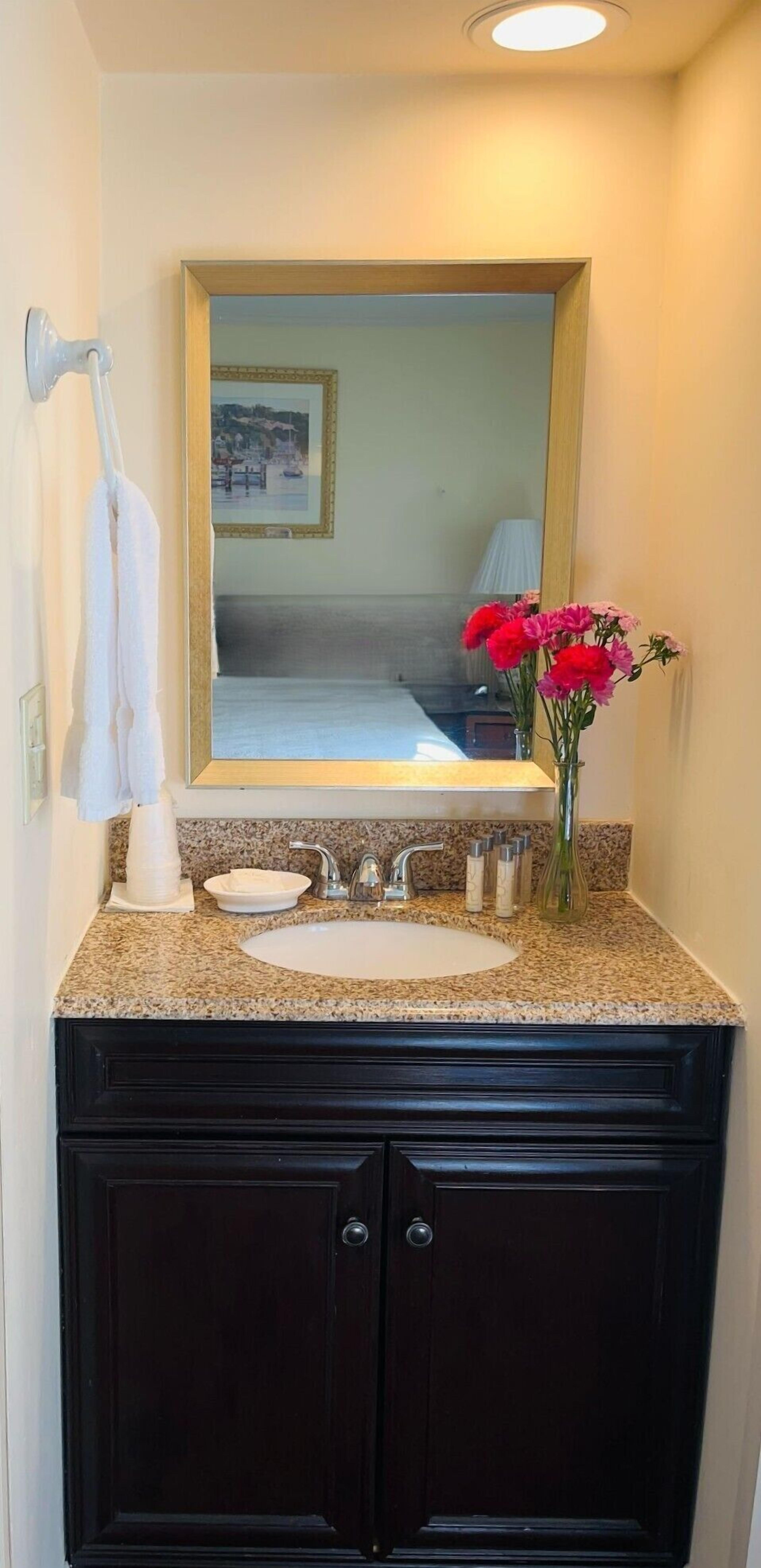 A bathroom with a granite countertop, a white sink, dark wooden cabinets, a mirror with a gold frame, and a vase with pink flowers.