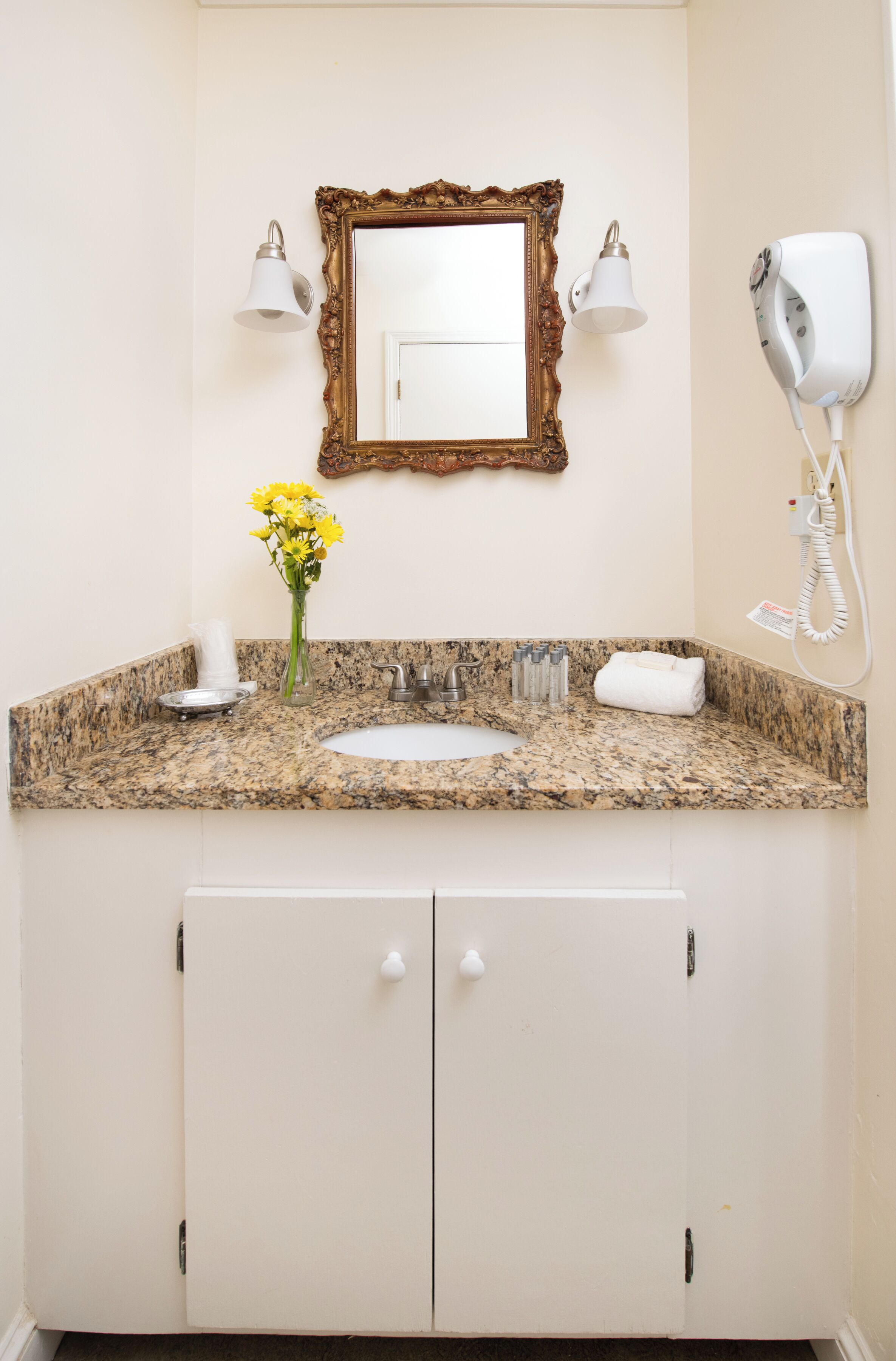 A bathroom with a granite countertop, a white sink, a mirror with a decorative frame, a hairdryer, and a vase with yellow flowers.