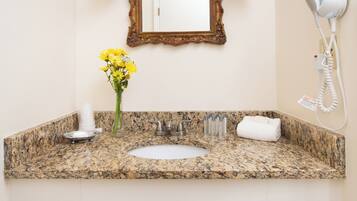A bathroom with a granite countertop, a white sink, a mirror with a decorative frame, a hairdryer, and a vase with yellow flowers.