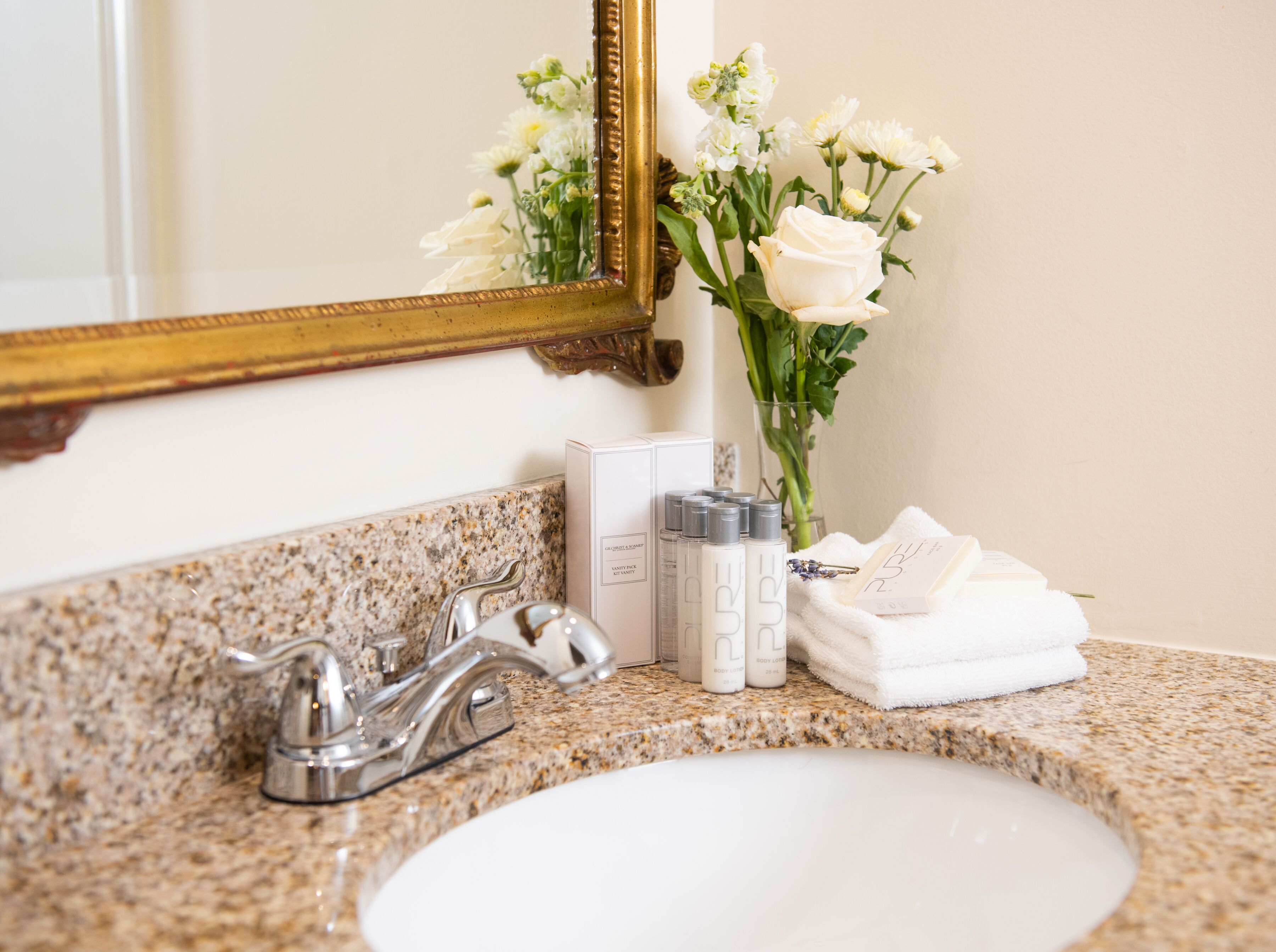 A bathroom vanity with a granite countertop, a white sink, a gold-framed mirror, and toiletries including body lotion bottles and a white towel.