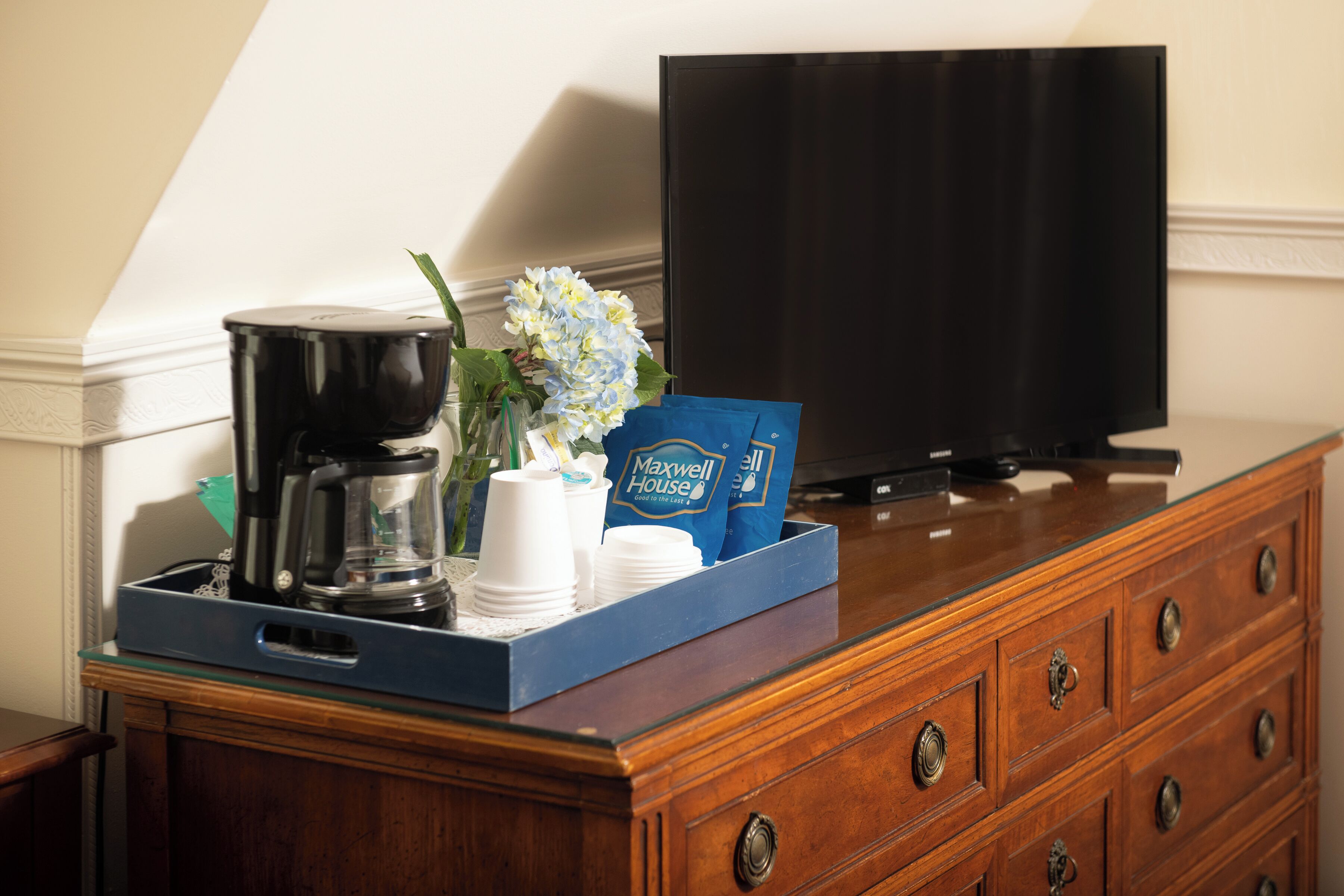 A wooden dresser with a tray holding a coffee maker, cups, a vase with flowers, and Maxwell House coffee bags. A television is mounted on the wall behind the dresser.