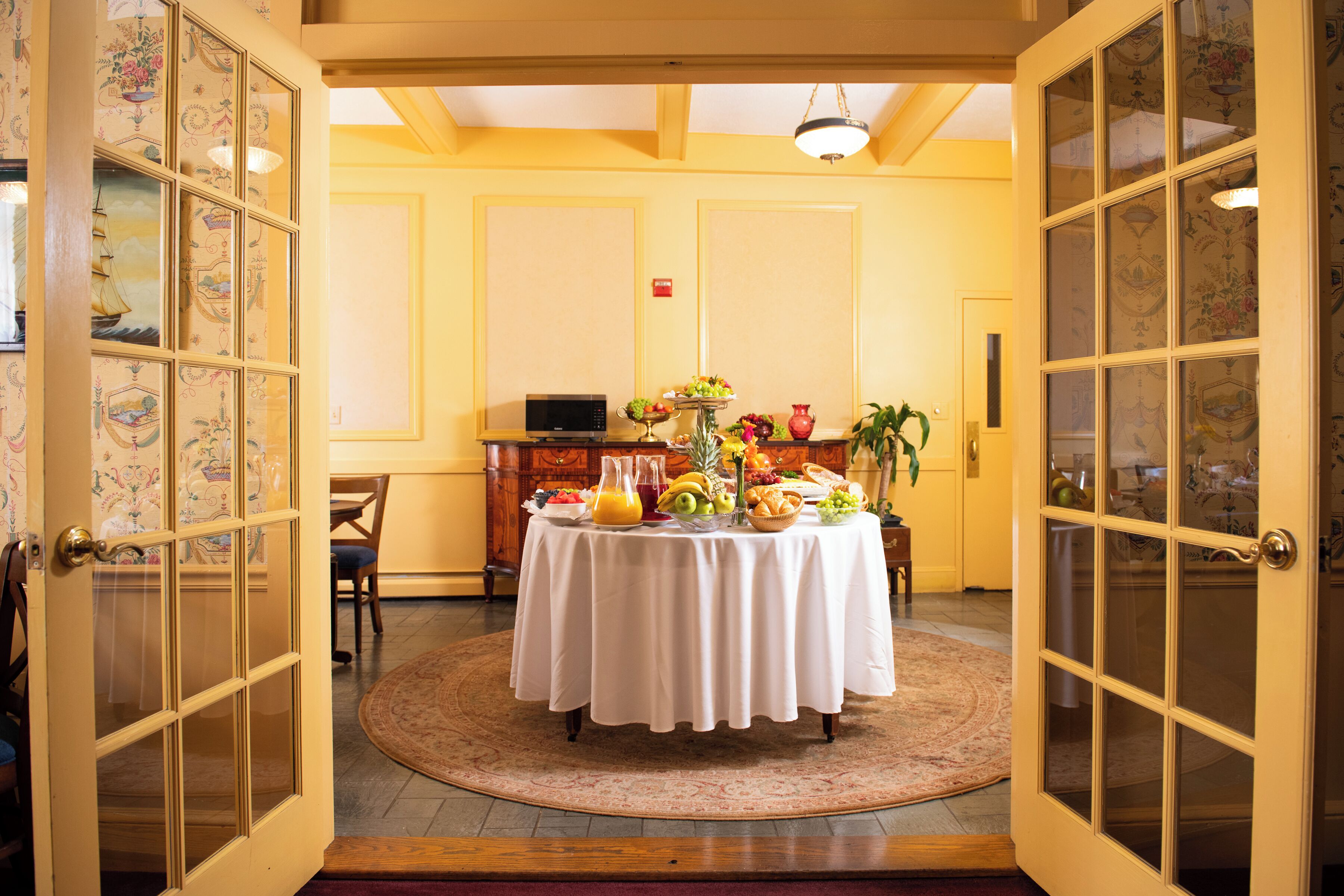 A room with a round table set for breakfast, a microwave, and a potted plant.