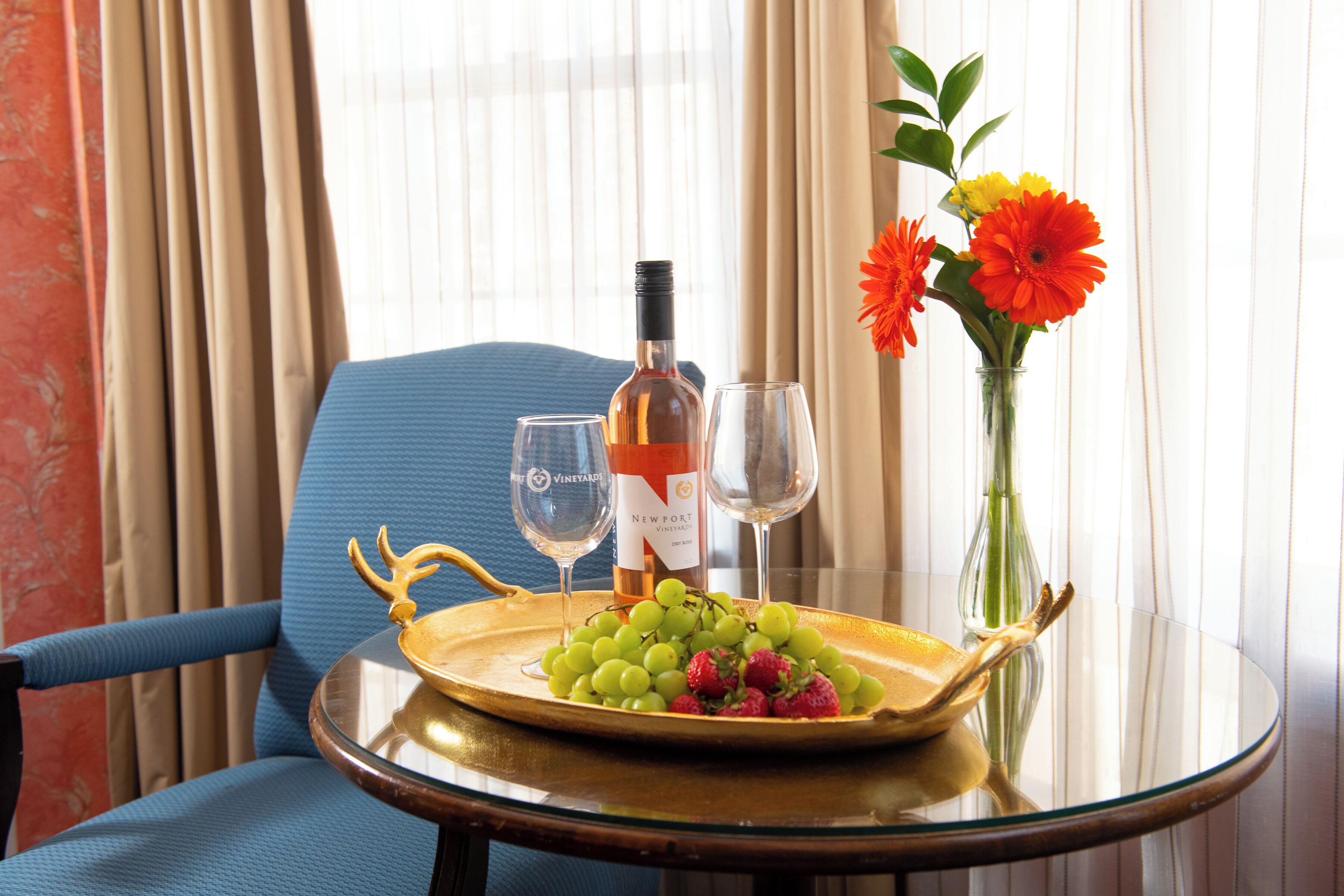 A table with a bottle of wine, two glasses, a tray with grapes and strawberries, and a vase with flowers.