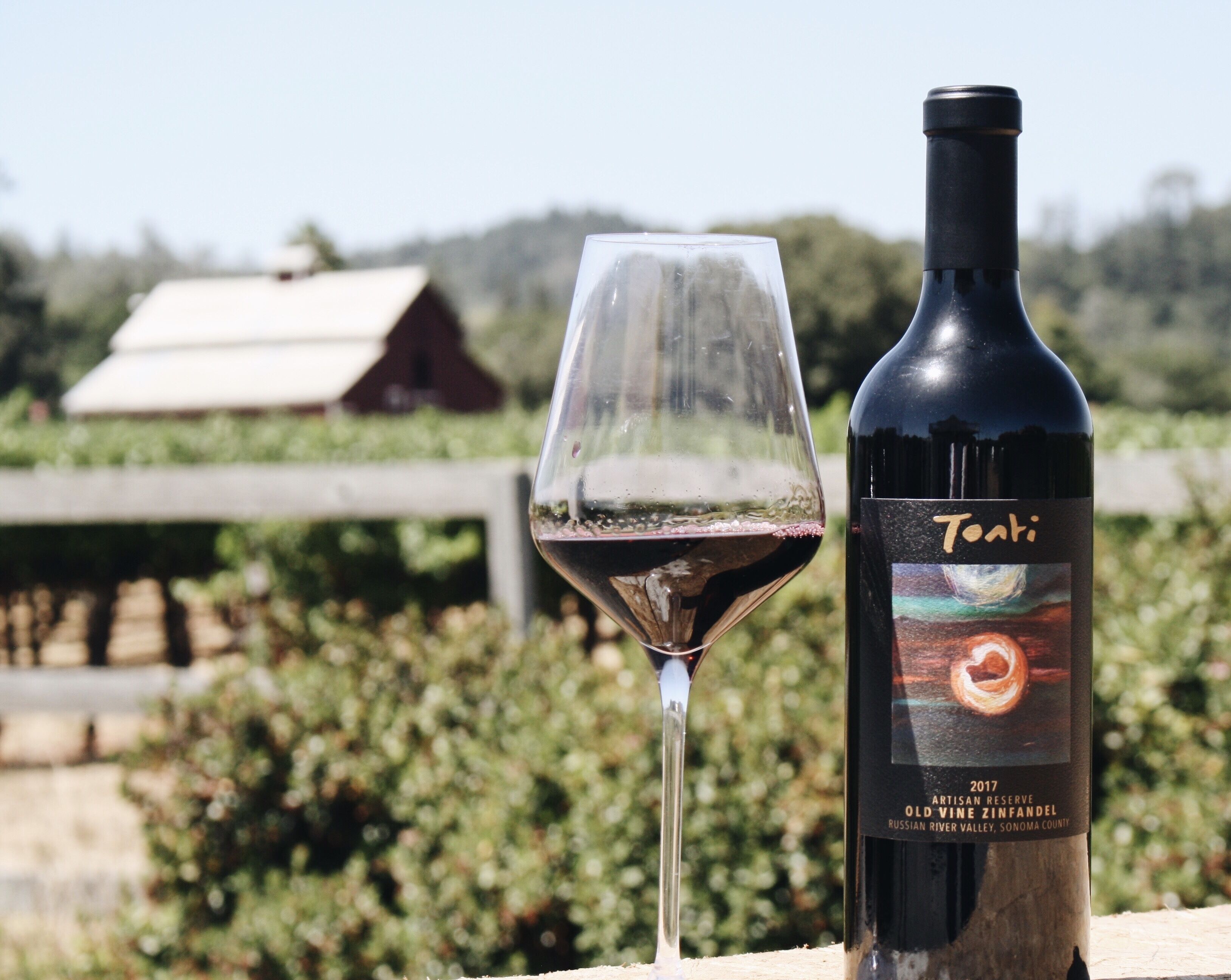 A bottle of Tokai wine and a glass of red wine on a wooden surface with a vineyard and a building in the background.