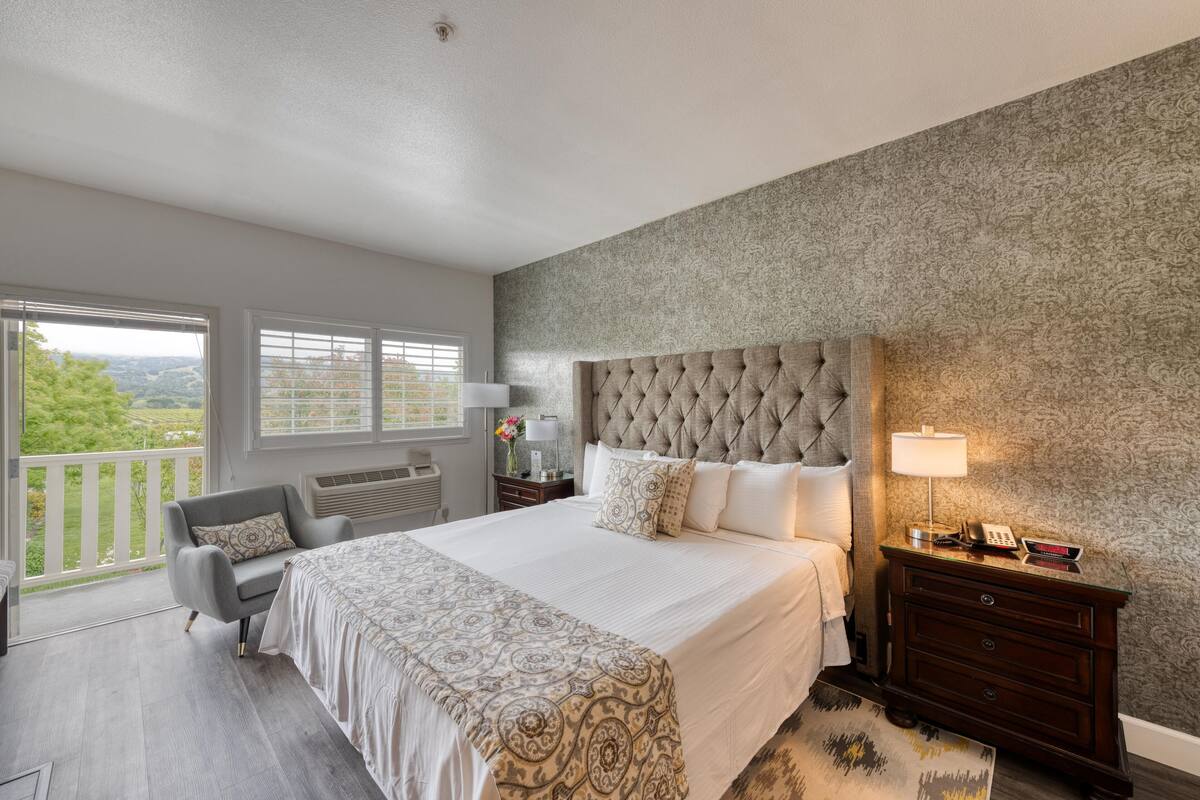 A bedroom with a large bed, a chair, a nightstand, and a view of greenery outside.