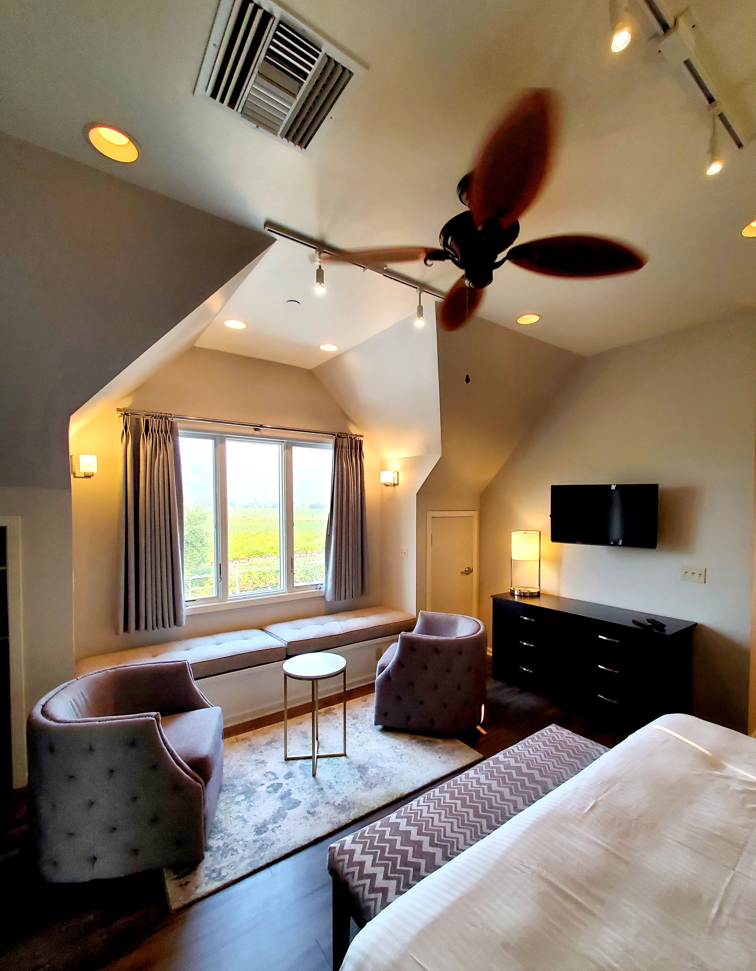 A bedroom with a bed, two armchairs, a small table, a ceiling fan, a TV, and a window with curtains.