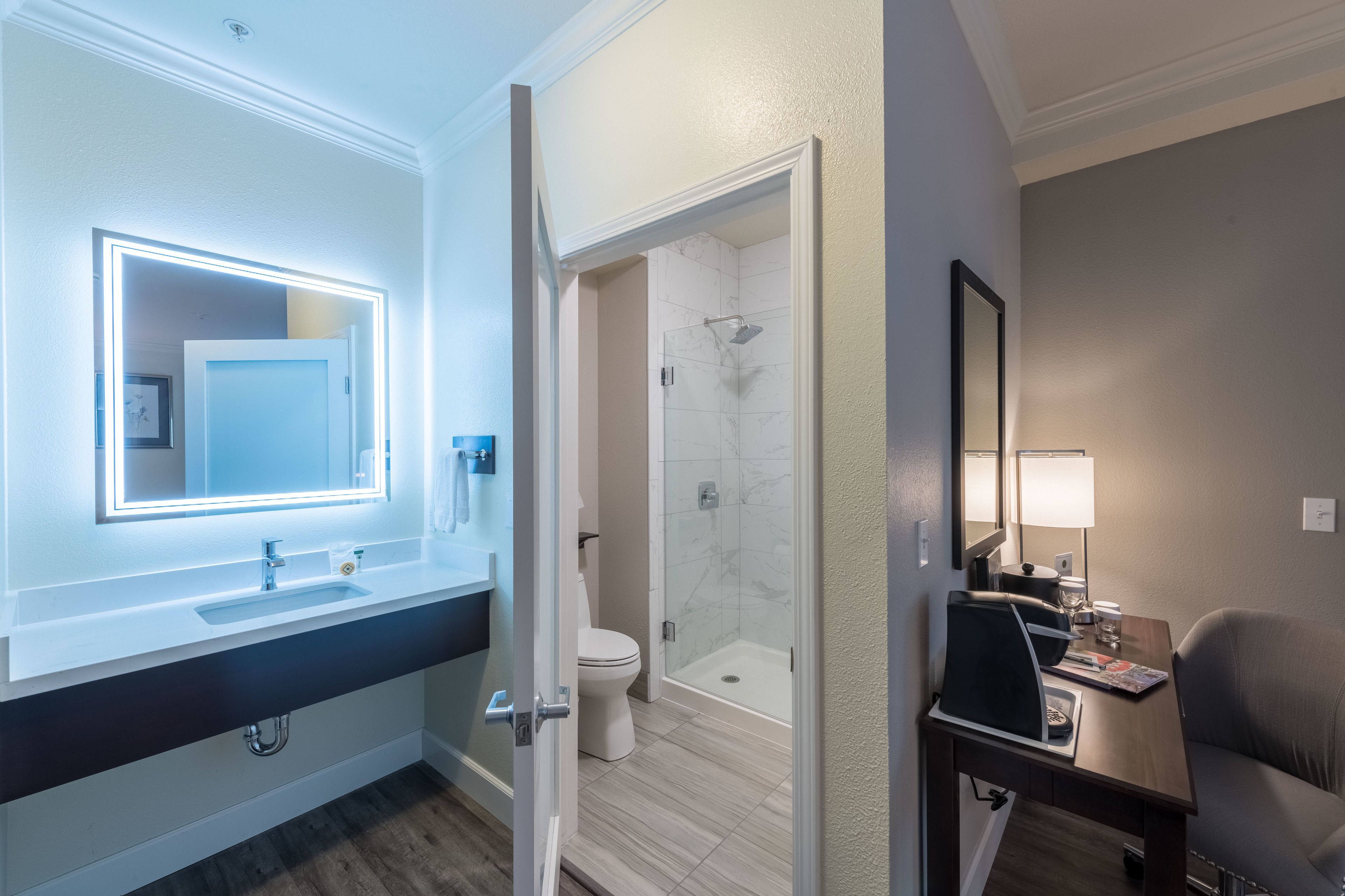 A modern bathroom with a vanity, a mirror with lights, a toilet, a shower, and a small table with a chair.