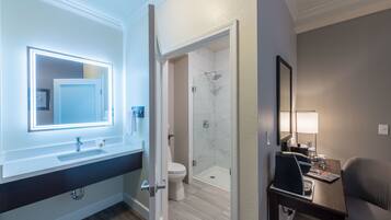 A modern bathroom with a vanity, a mirror with lights, a toilet, a shower, and a small table with a chair.