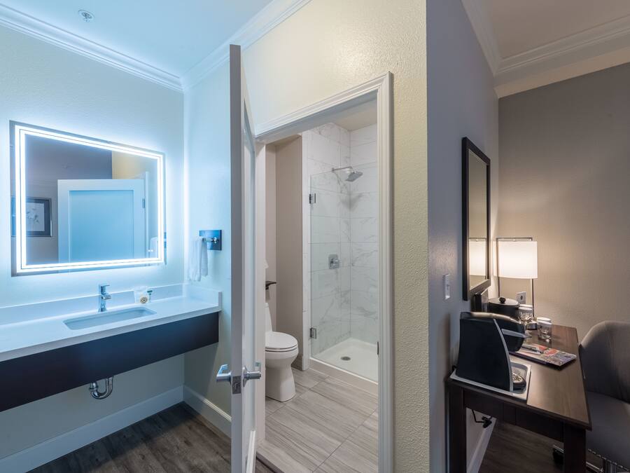 A modern bathroom with a vanity, a mirror with lights, a toilet, a shower, and a small table with a chair.