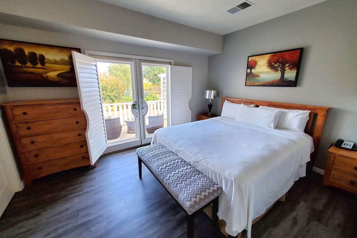 A bedroom with a large bed, a wooden dresser, a bench, a nightstand with a lamp, and a painting on the wall.