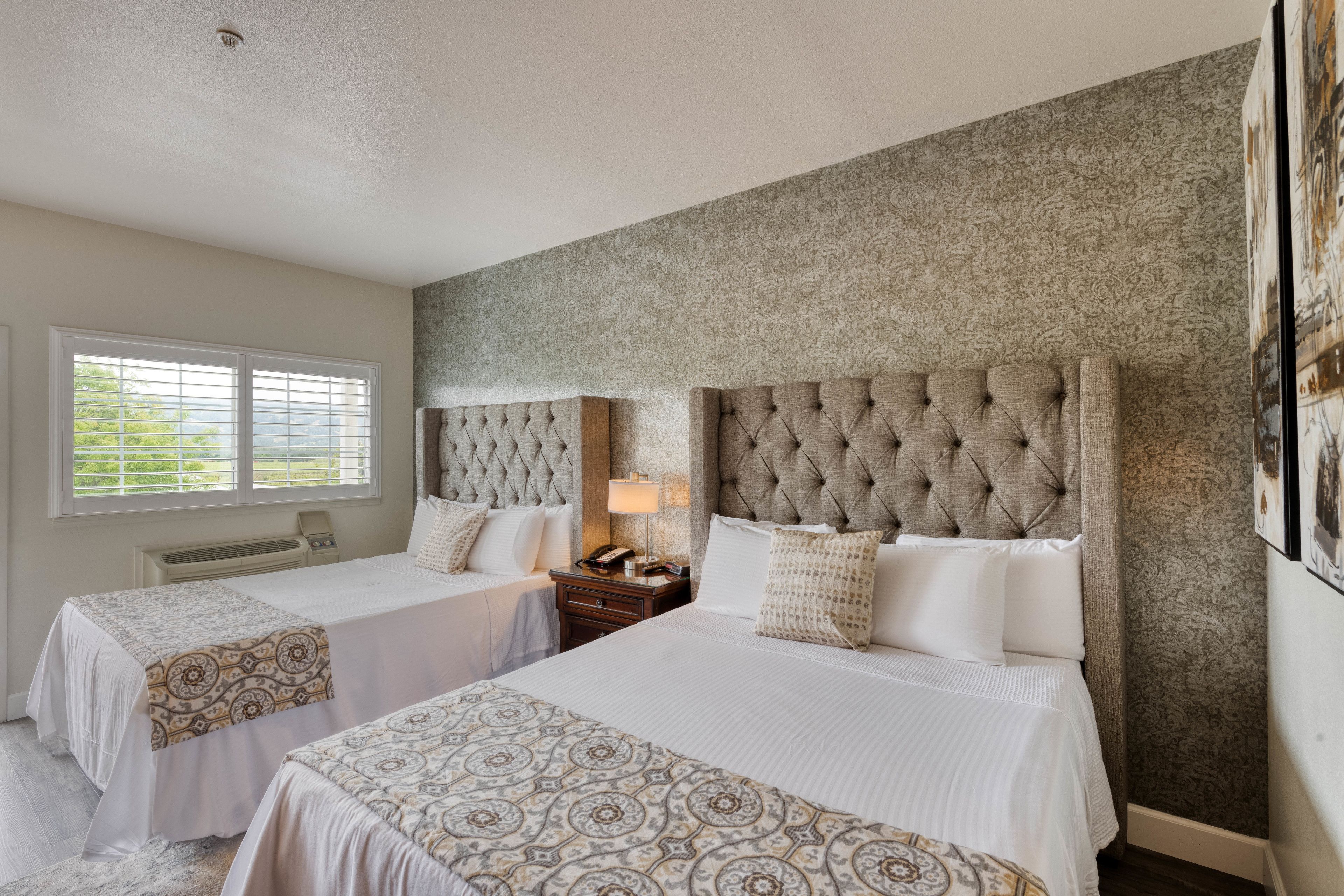 Habitación de hotel con dos camas, un ventanal con persianas, aire acondicionado de pared y papel tapiz estampado.