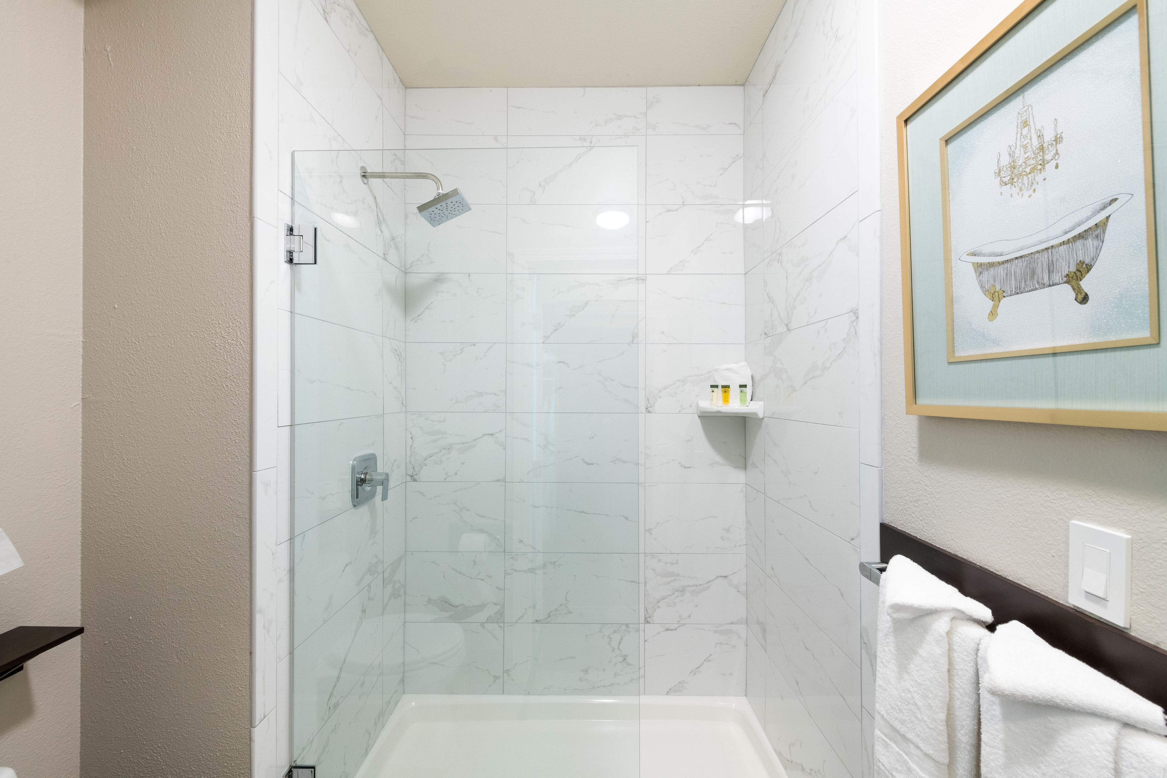 A modern bathroom with a glass-enclosed shower, marble tiles, a framed artwork of a bathtub, and a white towel hanging on the wall.