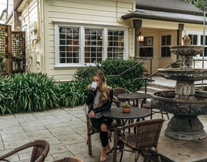 A woman sitting at an outdoor table with chairs, a fountain, and a building with large windows in the background.