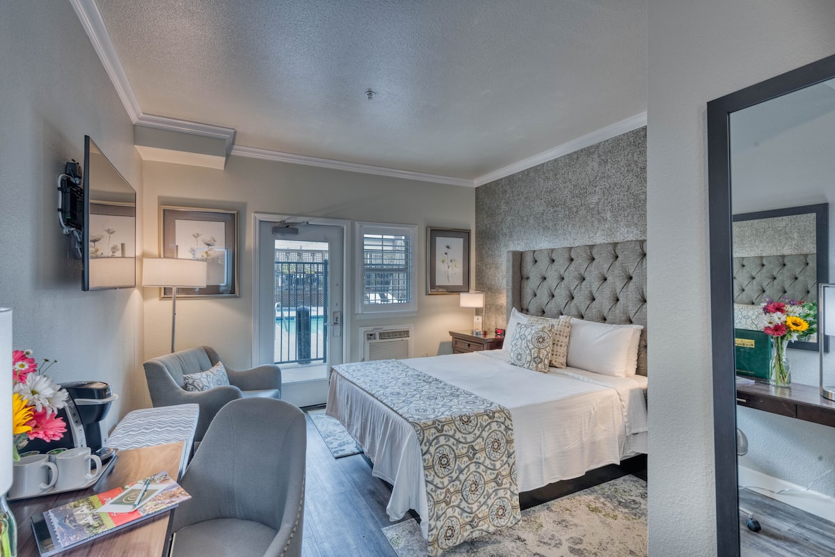 A hotel room with a large bed, a desk with a chair, a mirror, and a window with blinds.