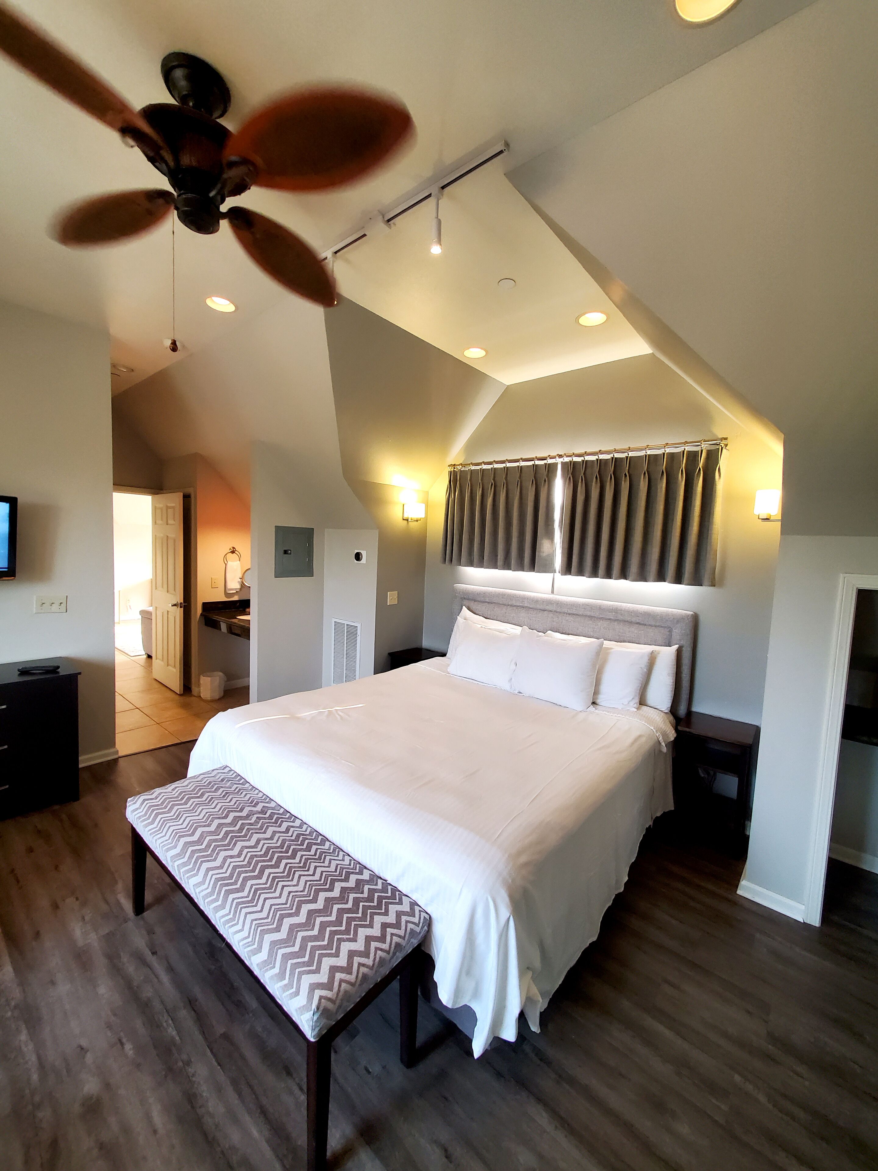 A bedroom with a large bed, a ceiling fan, a bedside table, a bench, and a view of the hallway.