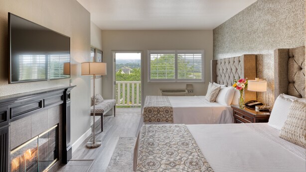 Quarto de hotel com lareira, duas camas, uma televisão grande e varanda com vista.
