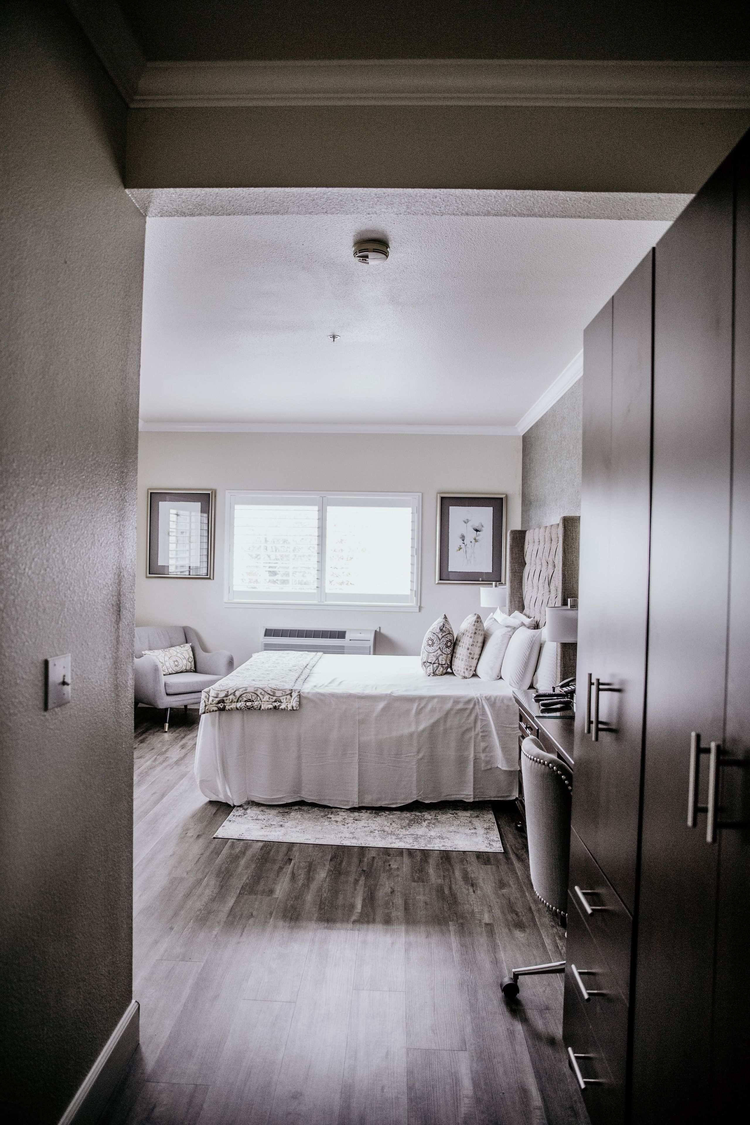 A modern bedroom with a large bed, a desk, and a chair. There are two windows and a radiator on the wall.