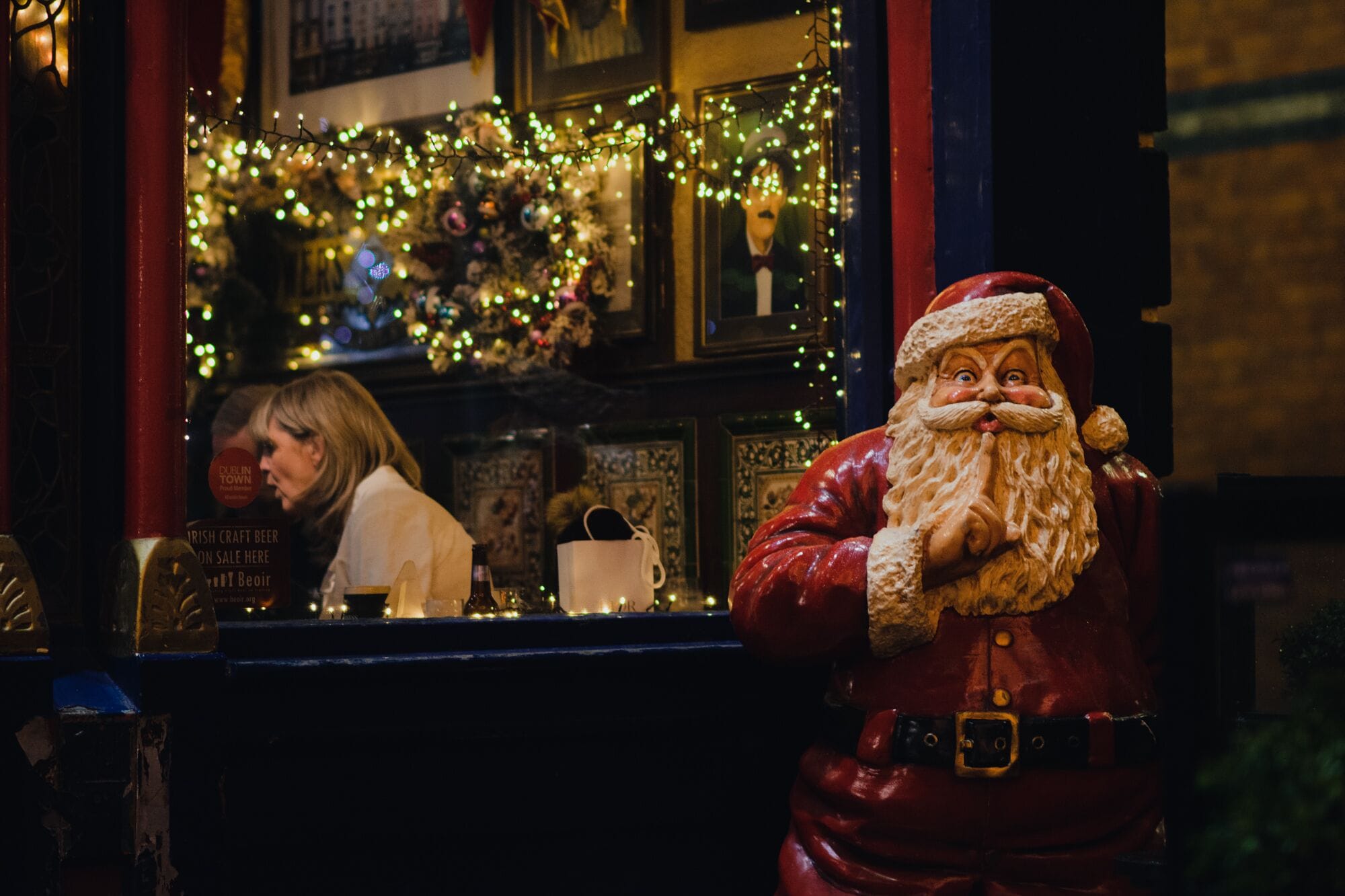 A statue of Santa Claus in front of a window with a wreath and lights.