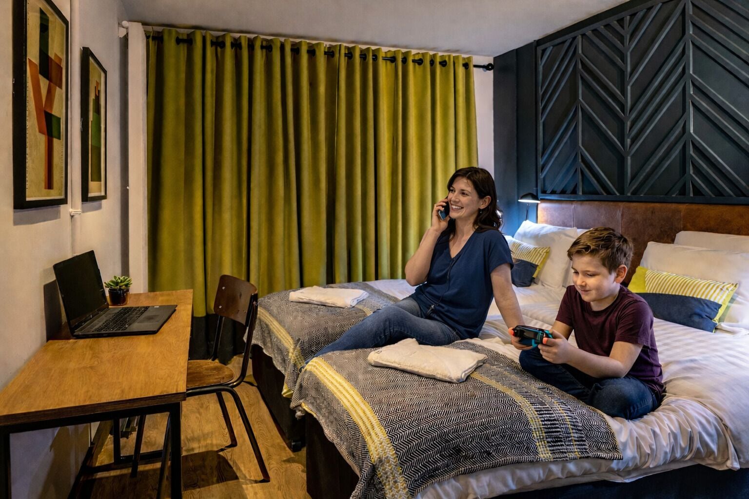 A hotel room with two beds, a desk, a laptop, and a green curtain.