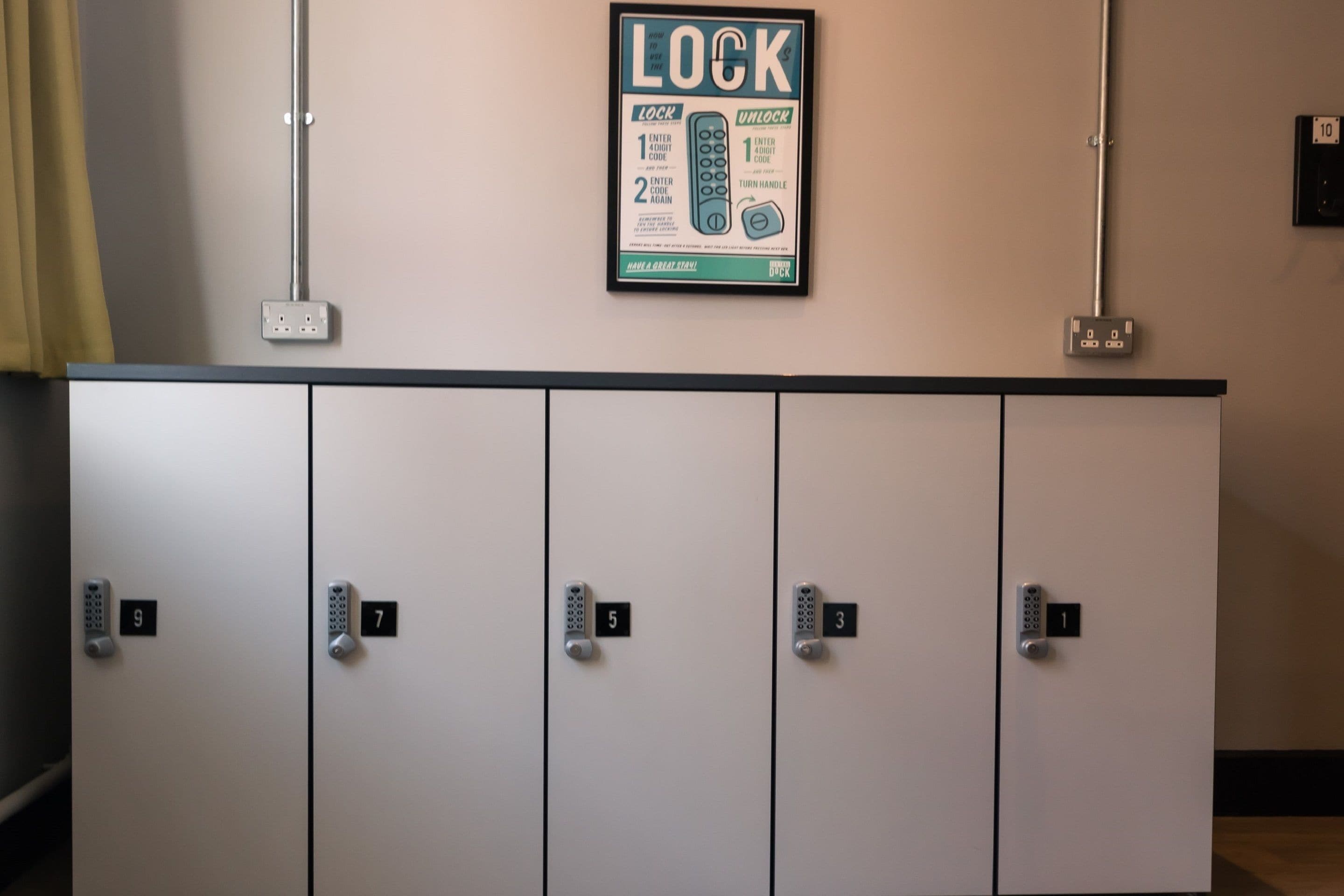 A row of lockers with numbered locks, a wall-mounted lock instruction poster, and electrical outlets above the lockers.