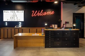 A reception desk with a neon 'Welcome' sign, a large lighted sign stating 'ALL YOU NEED IS LOVE AND A PASSPORT', and a counter with a laptop and a plant. - Latroupe Jacobs Inn (Dublin)
