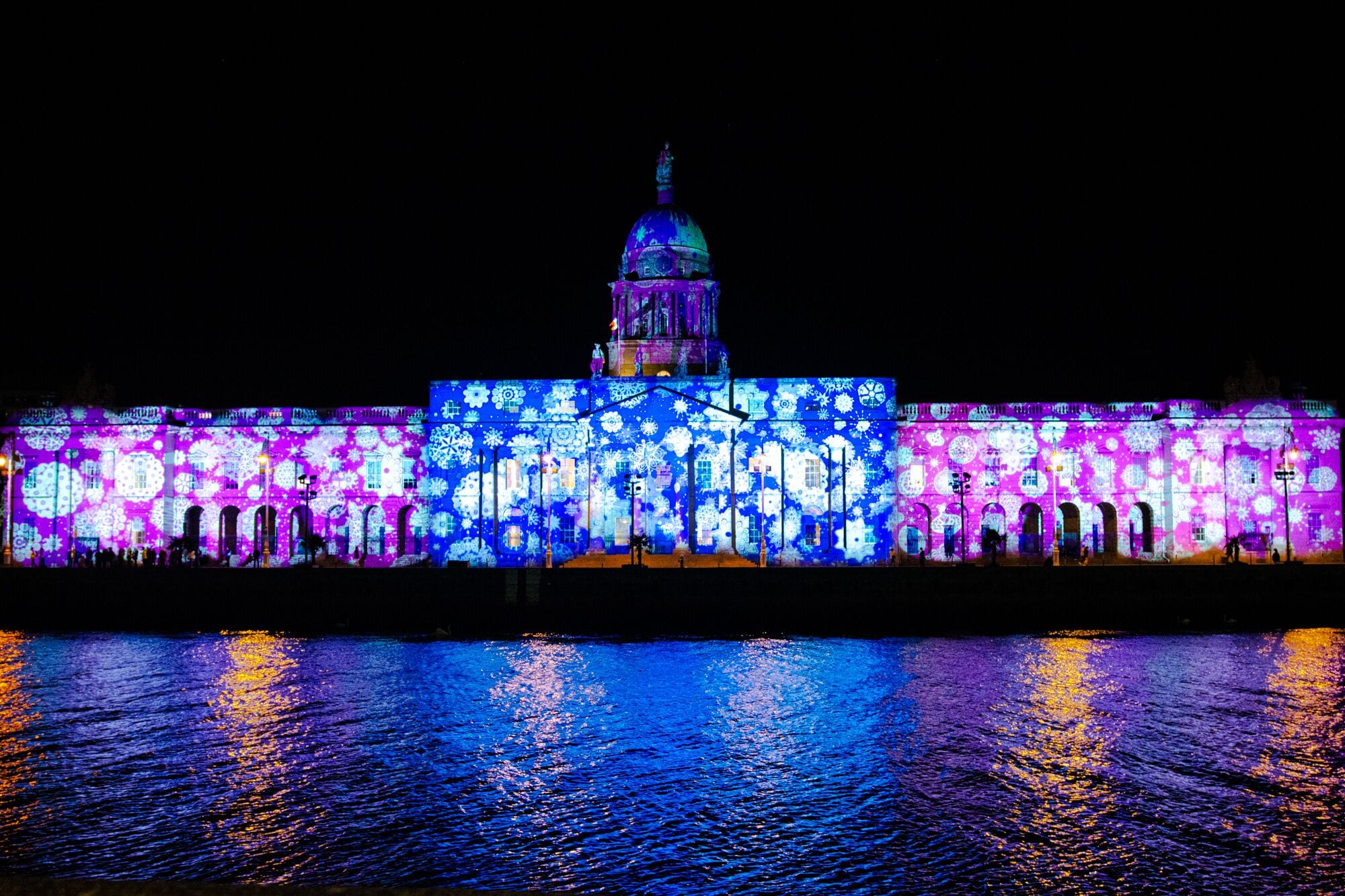A building with a large dome and arched windows, illuminated with colorful lights reflecting on the water.