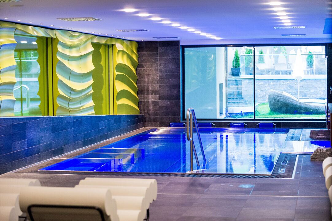A modern indoor swimming pool with clear glass walls, a blue floor, and a view of the outside.