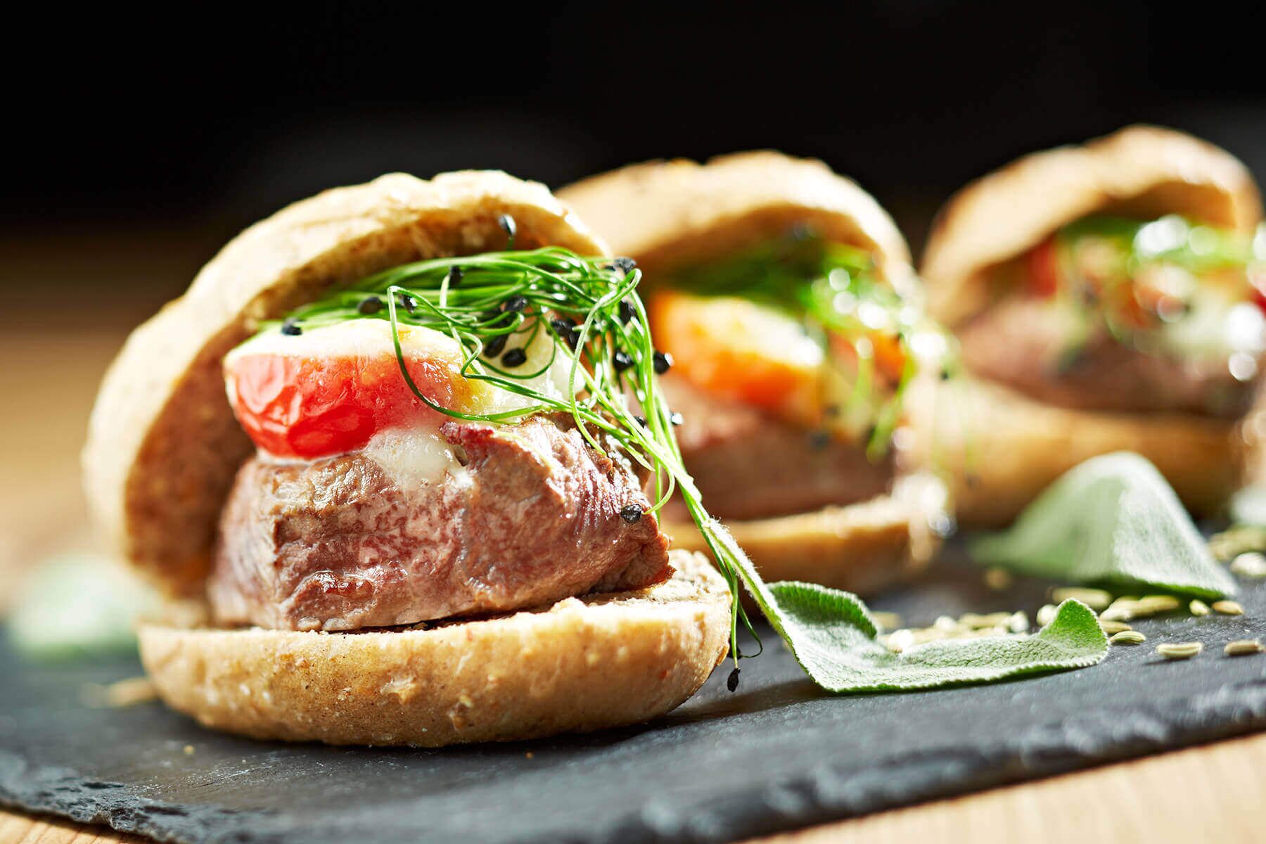 A sandwich with meat, tomato, and greens on a bun, placed on a slate board.