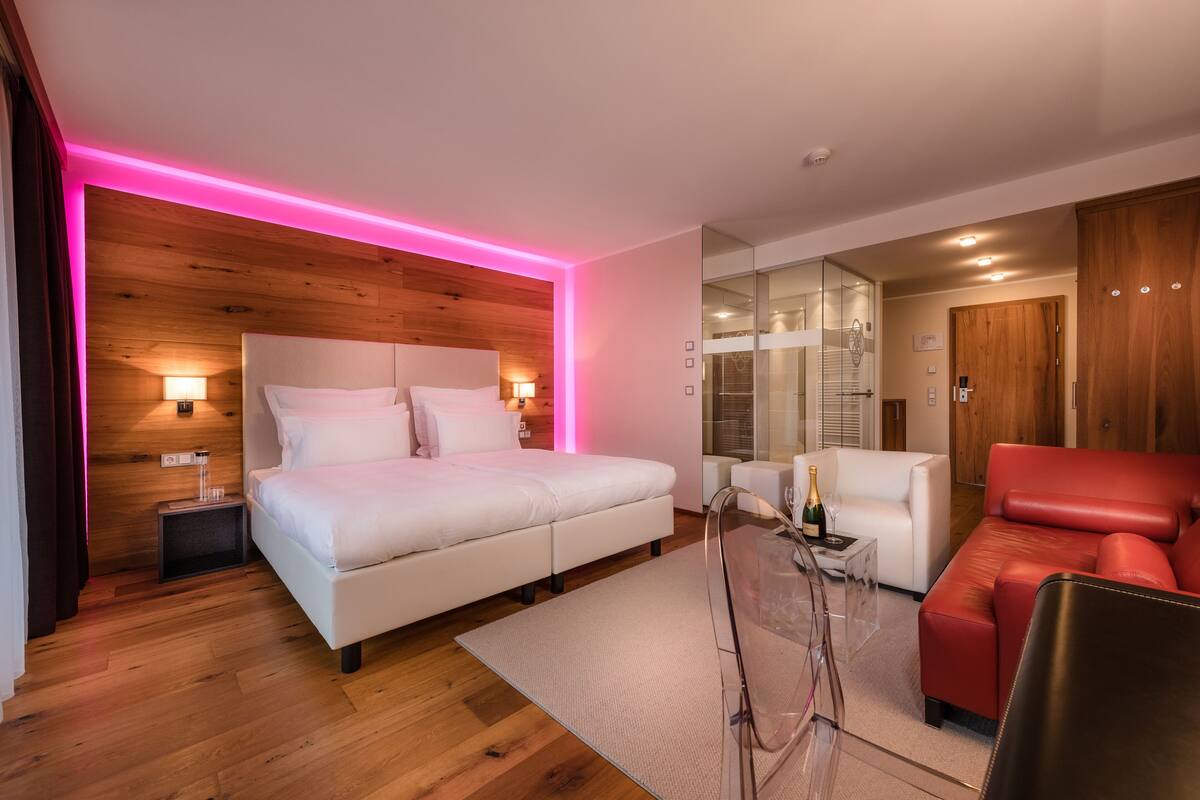 A modern hotel room with a wooden headboard, a white bed, a red leather sofa, a glass shower, and a wooden wardrobe.