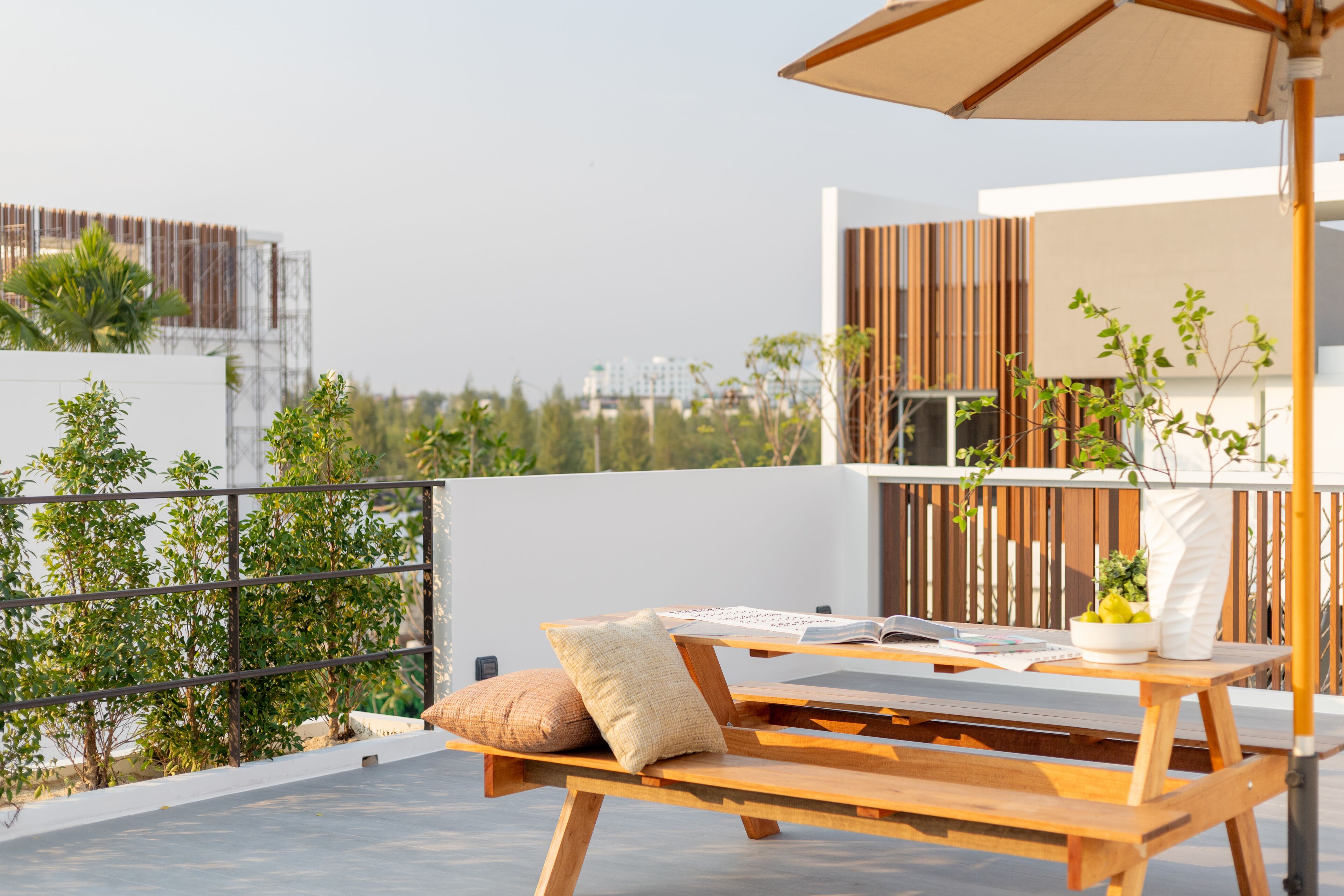 A wooden lounge chair with a cushion, a table with a book, and a bowl of fruit on a rooftop terrace with a view of buildings and trees.