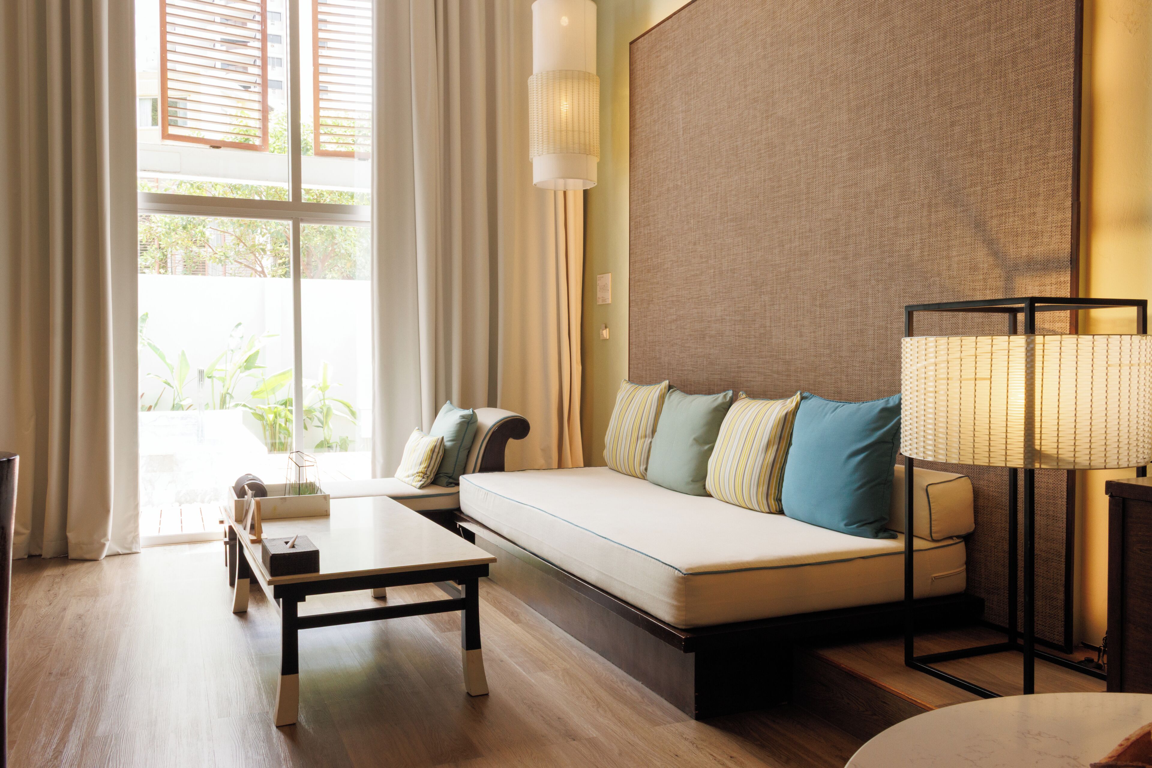 A modern hotel room with a sofa, a side table, a lamp, and a view of greenery outside.