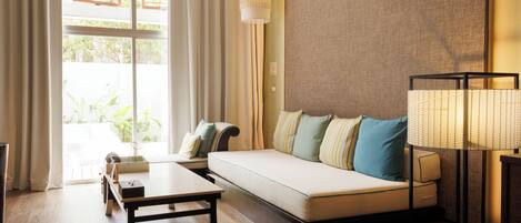 A modern hotel room with a sofa, a side table, a lamp, and a view of greenery outside.