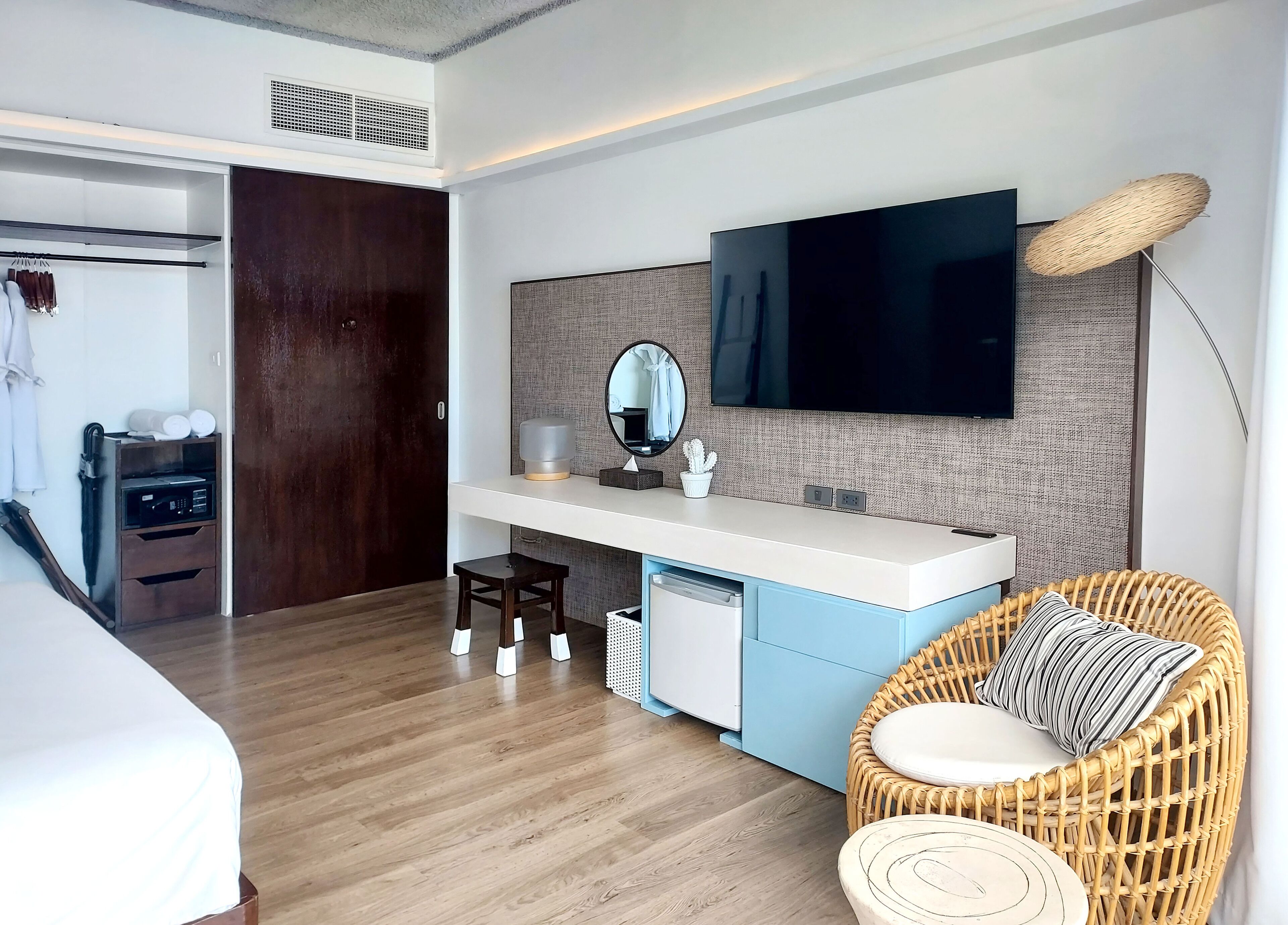 Quarto de hotel moderno com piso de madeira, uma cama, um guarda-roupa, uma toucador com espelho, televisão e uma cadeira de vime.