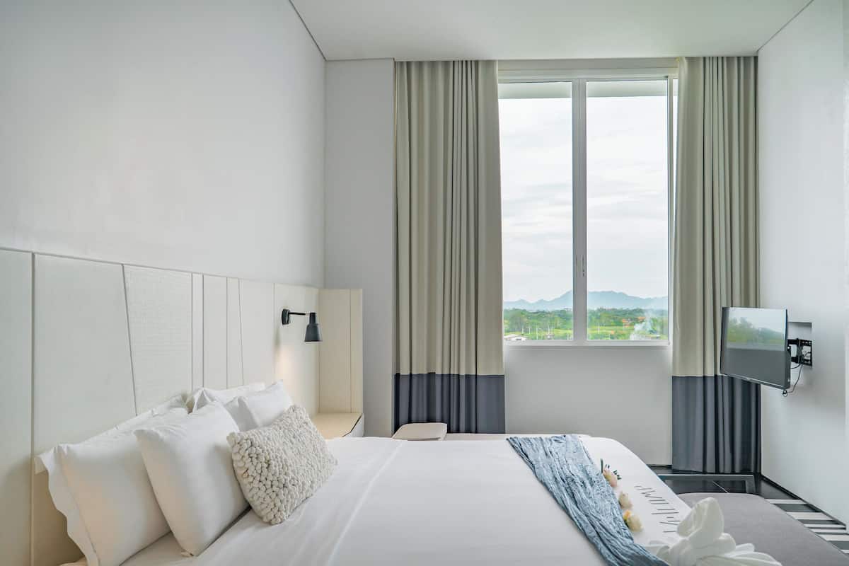 Ein Hotelzimmer mit Bett, Nachttisch, Lampe, Fernseher und einem Fenster mit Vorhängen.