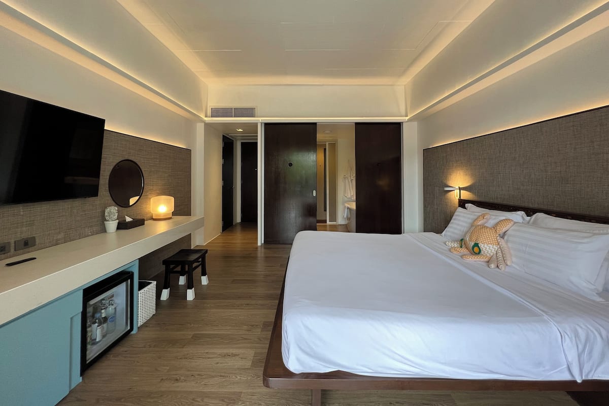 Ein modernes Hotelzimmer mit einem großen Bett, einem flachbildfernseher an der Wand, einem Nachttisch mit Lampe sowie einer kleinen Sitzecke mit einem Sessel und einem Hocker.