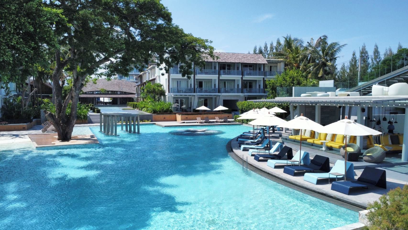 Un área de piscina con sillones de descanso y sombrillas, rodeada de edificios y árboles.