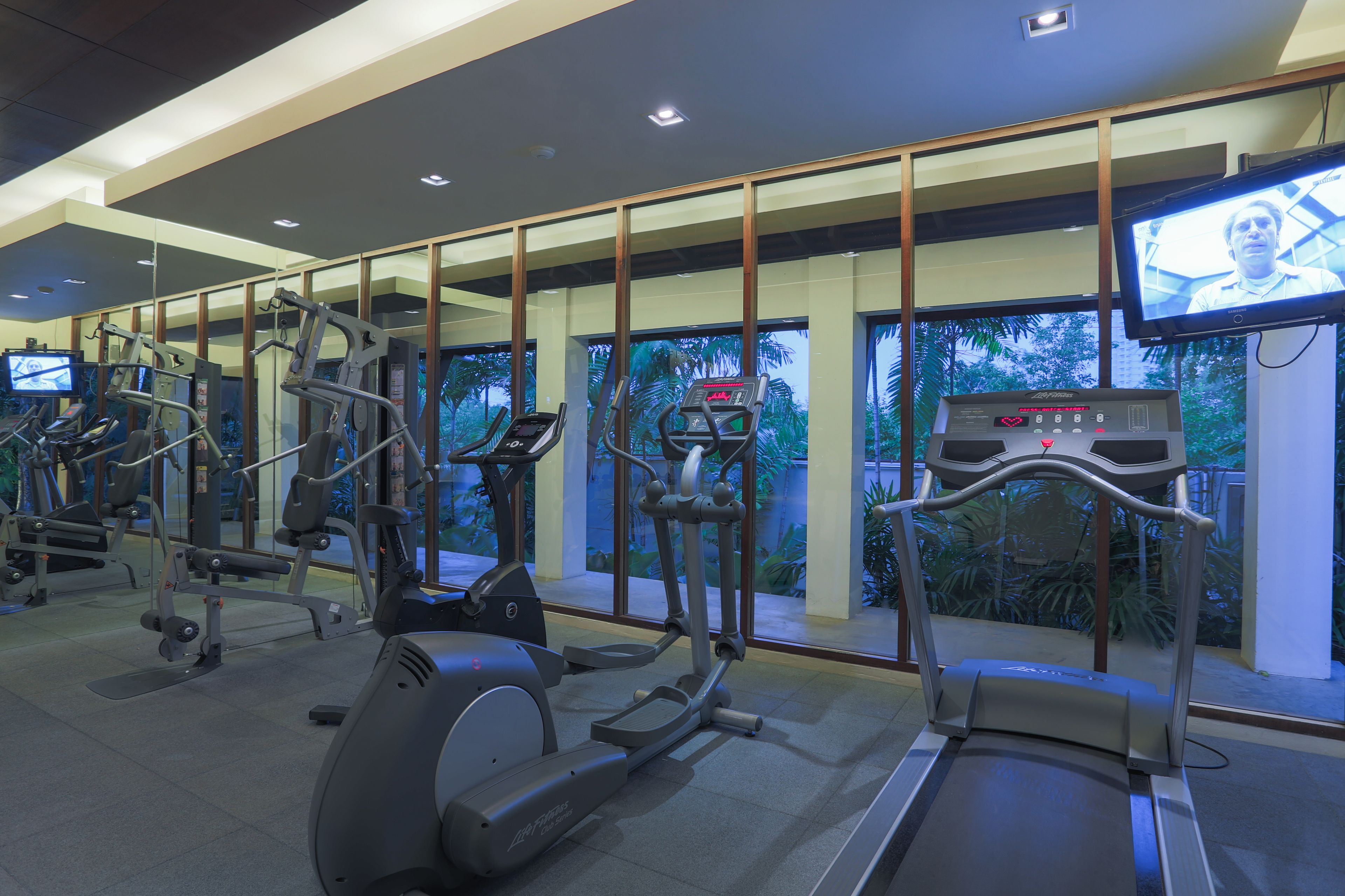 Un gimnasio con diversas máquinas de ejercicio, una pantalla de televisión y grandes ventanales con vistas al verdor.