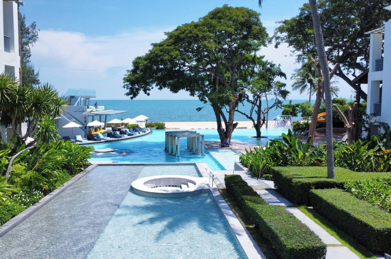 A resort pool area with a clear view of the ocean, surrounded by palm trees and lounge chairs.