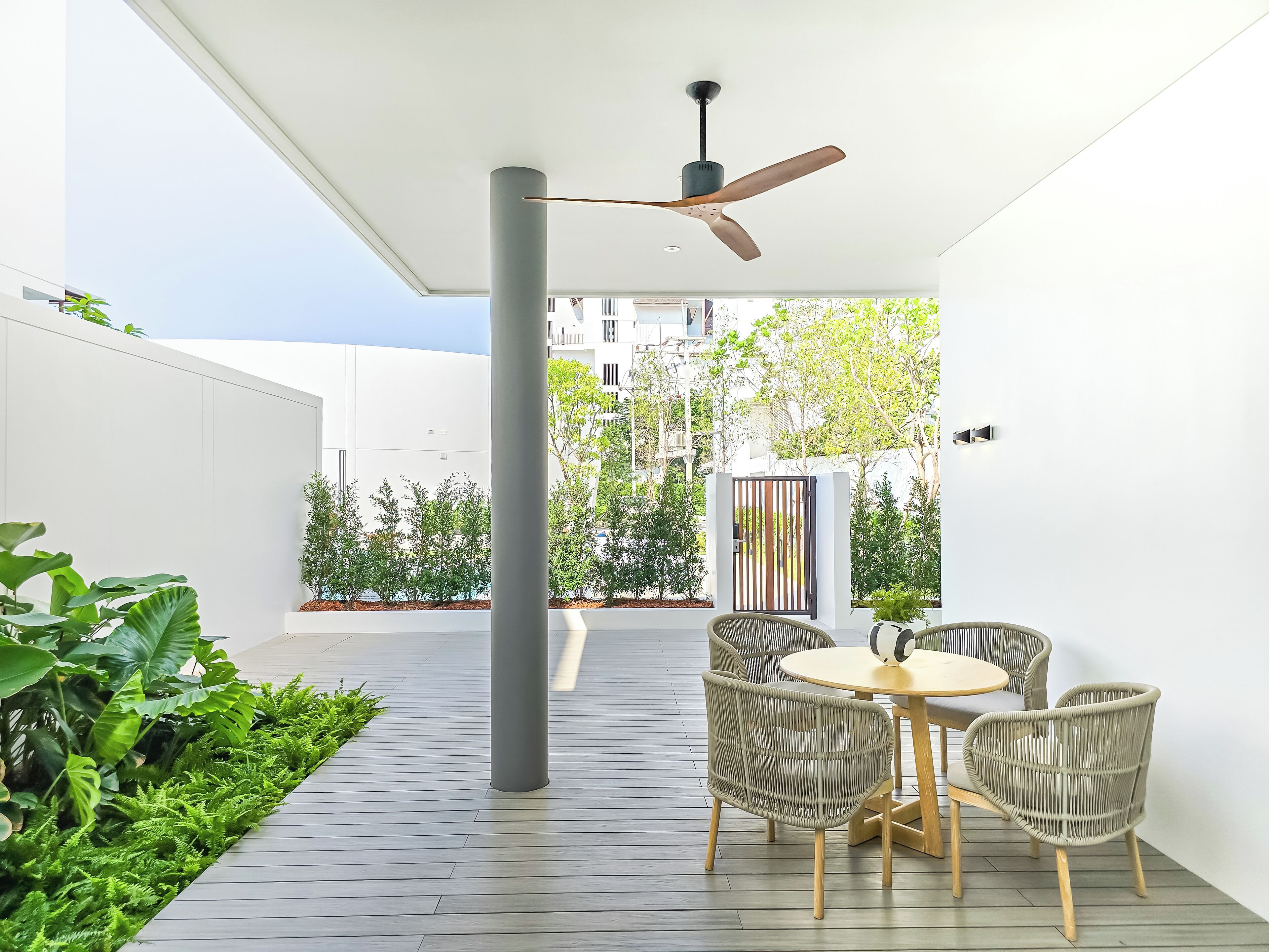 Une terrasse extérieure moderne, dotée d'une table ronde et de chaises en osier, d'un ventilateur de plafond et offrant une vue sur un quartier résidentiel.