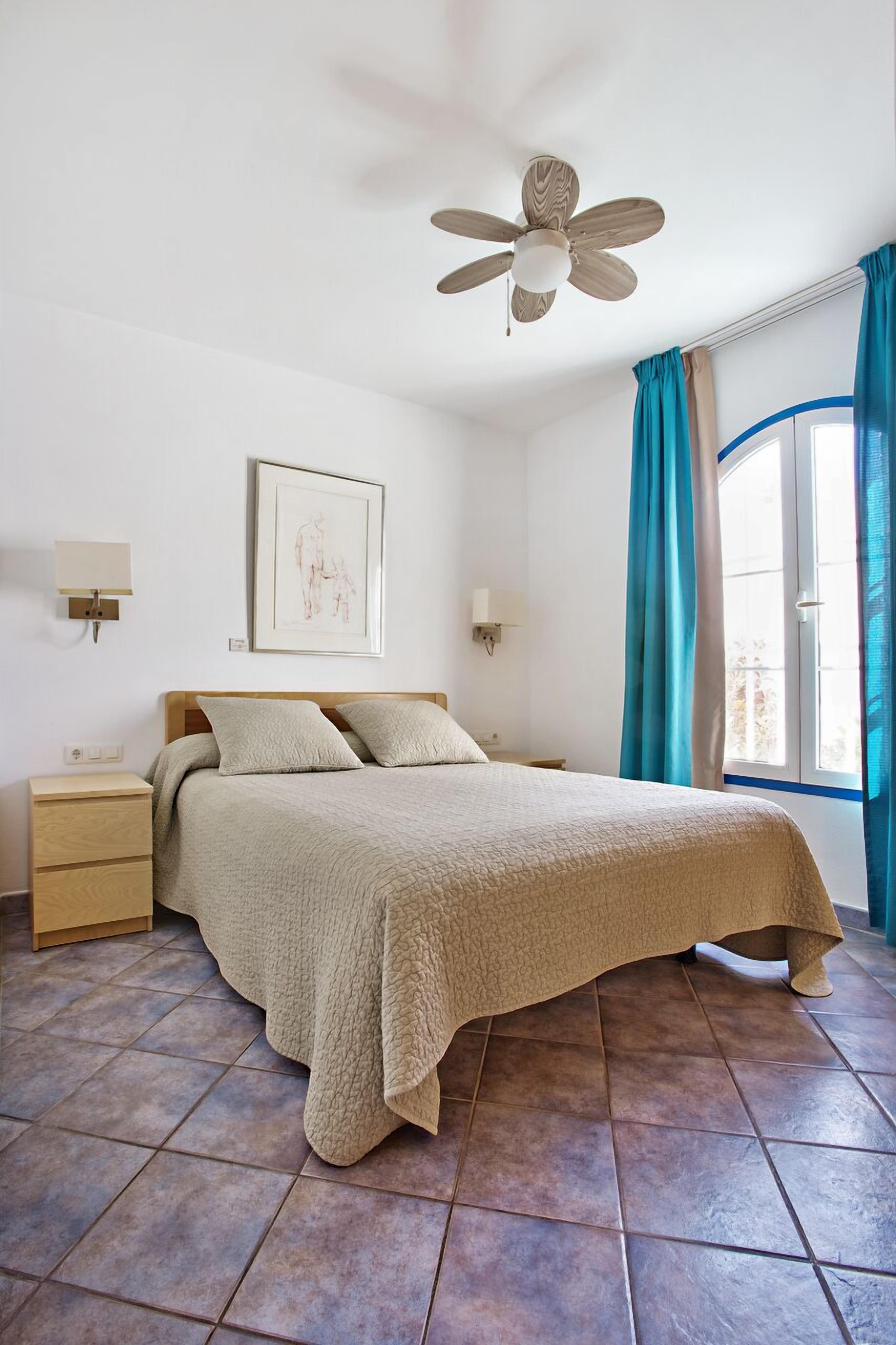 A bedroom with a bed, a nightstand, a ceiling fan, a window with curtains, and a wall-mounted light.