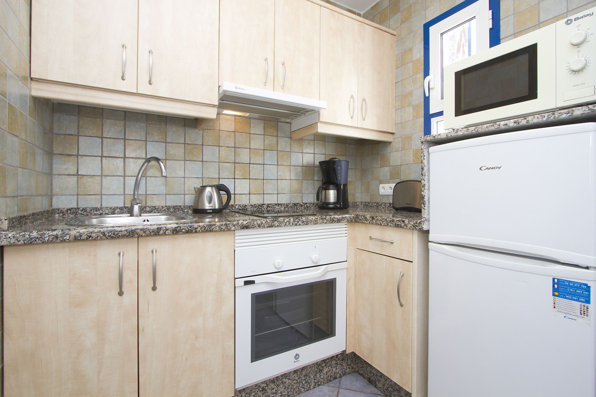 Una cocina compacta con armarios de madera clara, horno empotrado, microondas y cafetera.