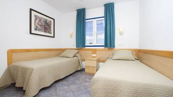 Une chambre avec deux lits simples, une fenêtre avec des rideaux, un luminaire fixé au mur et un tableau encadré.