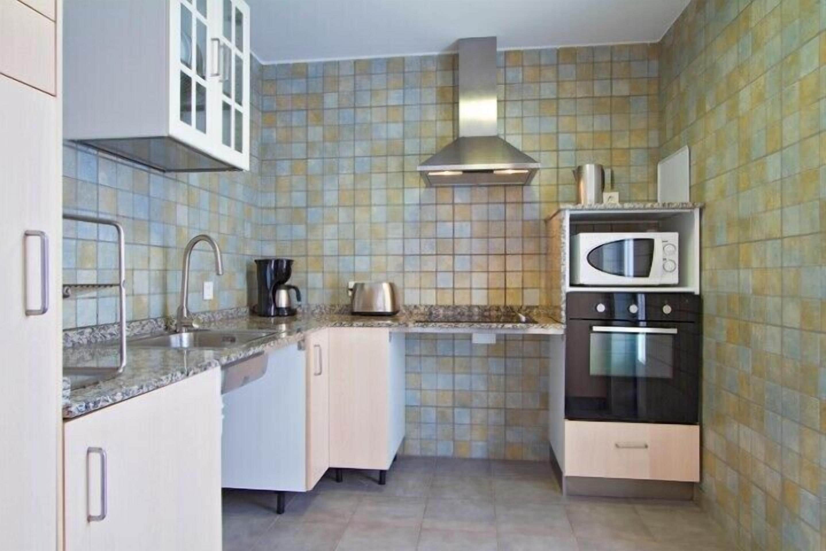 A compact kitchen with a tiled backsplash, a stainless steel range hood, a microwave, and a kettle on the countertop.