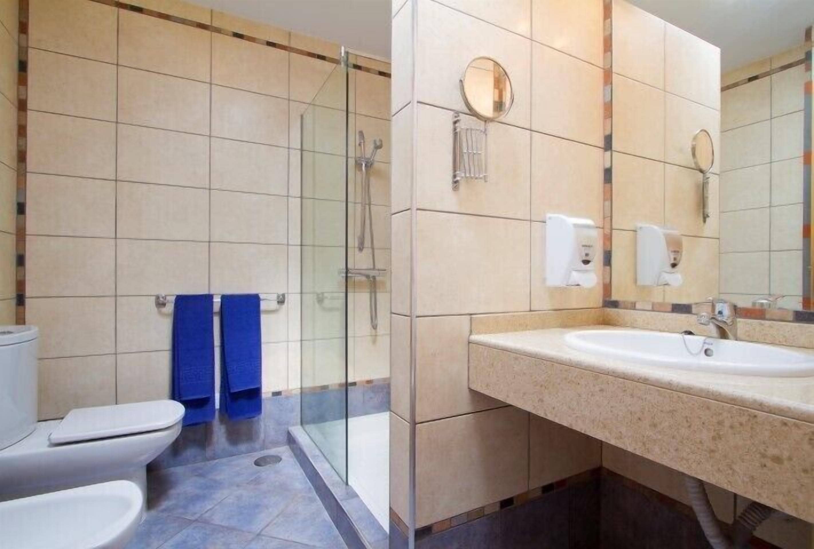 A modern bathroom with a glass-enclosed shower, a white sink, and two blue towels on the floor.
