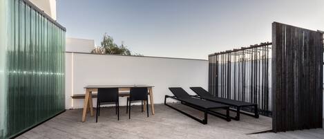 Terraza en la azotea con una mesa, sillas, dos tumbonas y una barandilla de cristal.