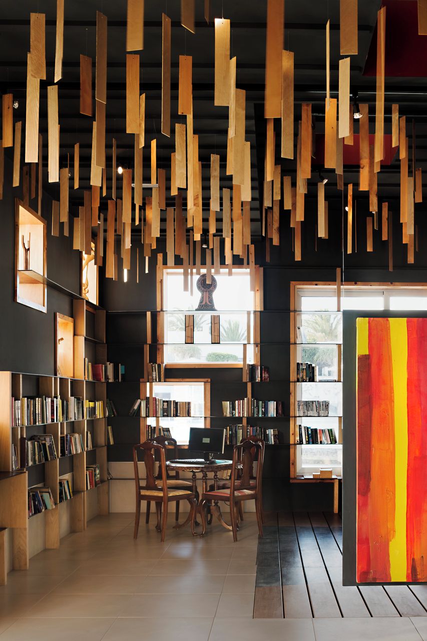 A library interior with bookshelves, a dining table, chairs, and hanging wooden elements.