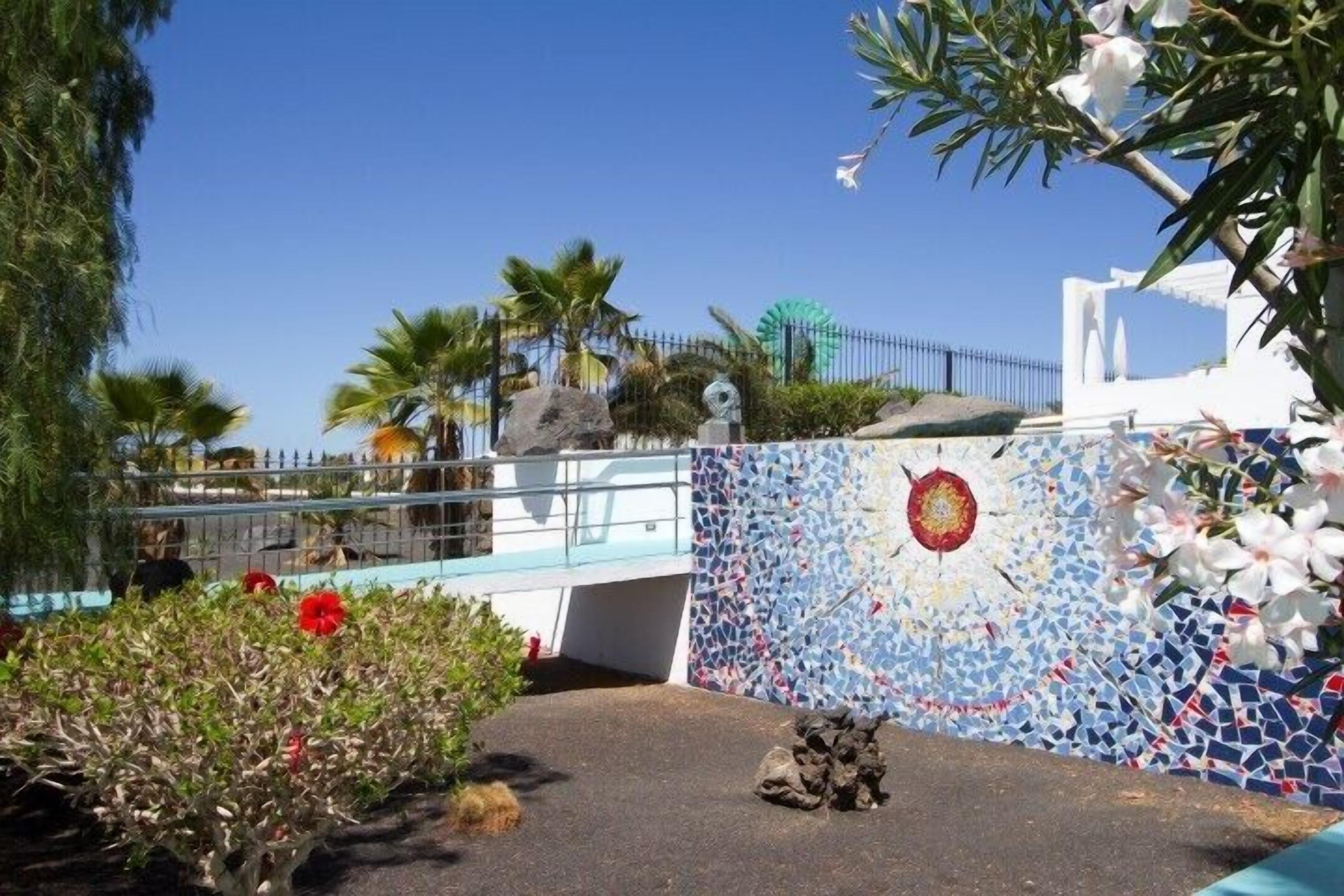 Una pared de mosaicos con un diseño circular, palmeras y un edificio blanco con techo azul.