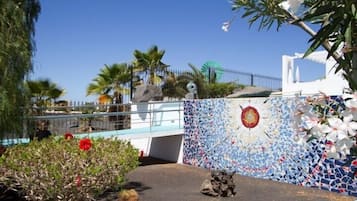 Una pared de mosaicos con un diseño circular, palmeras y un edificio blanco con techo azul.