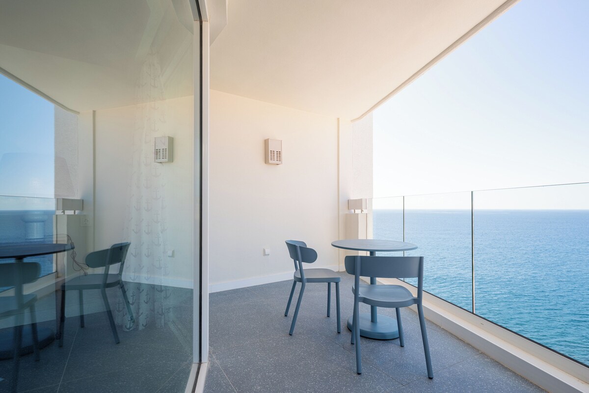A balcony with a glass partition, a round table, two chairs, and a view of the sea.